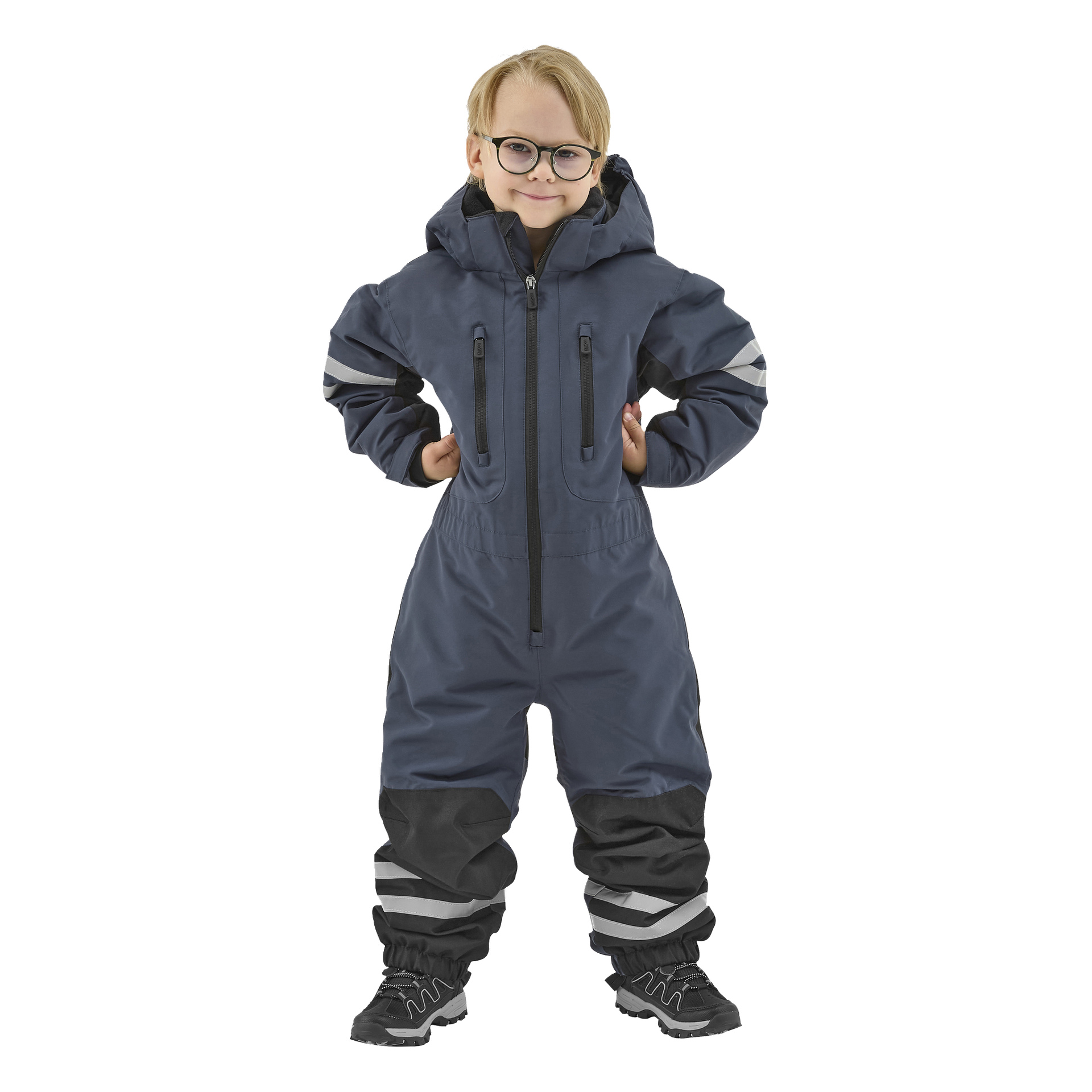 Ett litet barn med glasögon står vänd mot kameran i en mörkblå Kayoba Outdoor Dundret hooded snöoverall med reflekterande ränder på armar och ben, tillsammans med svarta stövlar. Barnets händer är placerade på höfterna och de har ett svagt leende.