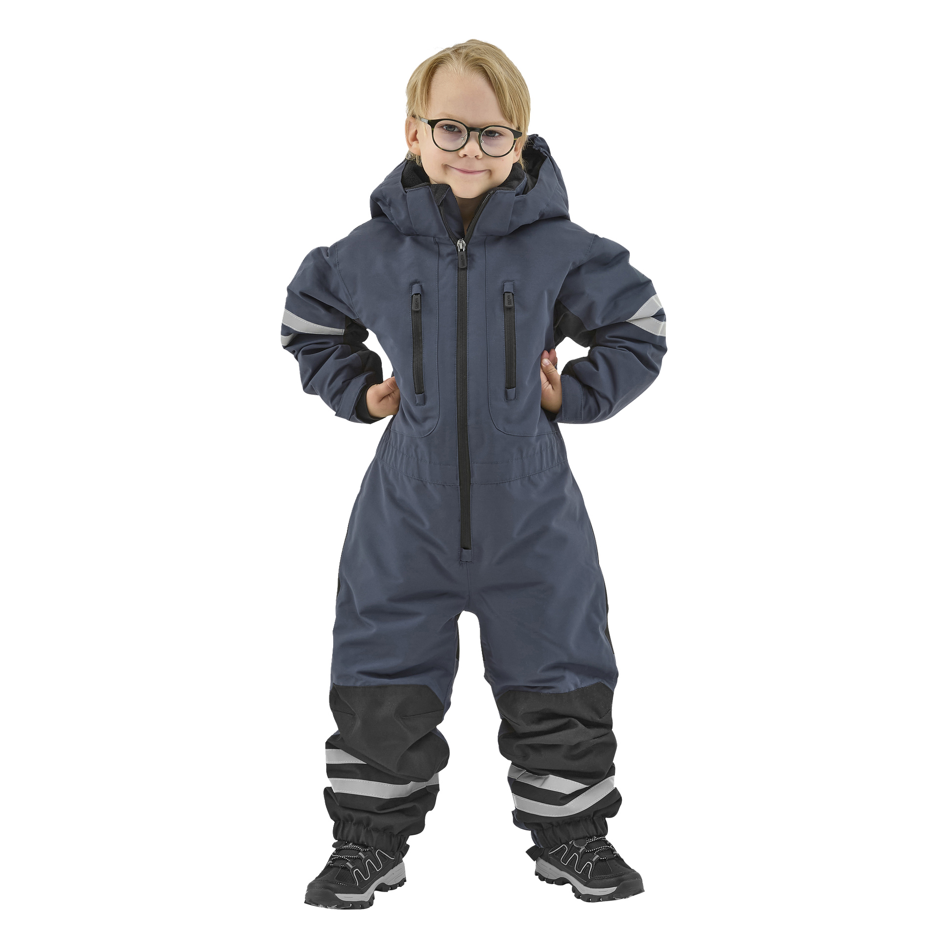 Ett leende barn med glasögon står självsäkert, händerna på höfterna, iklädd en mörkblå Kayoba Outdoor Dundret hooded snöoverall med reflekterande ränder och förstärkta knän, tillsammans med svarta skor.
