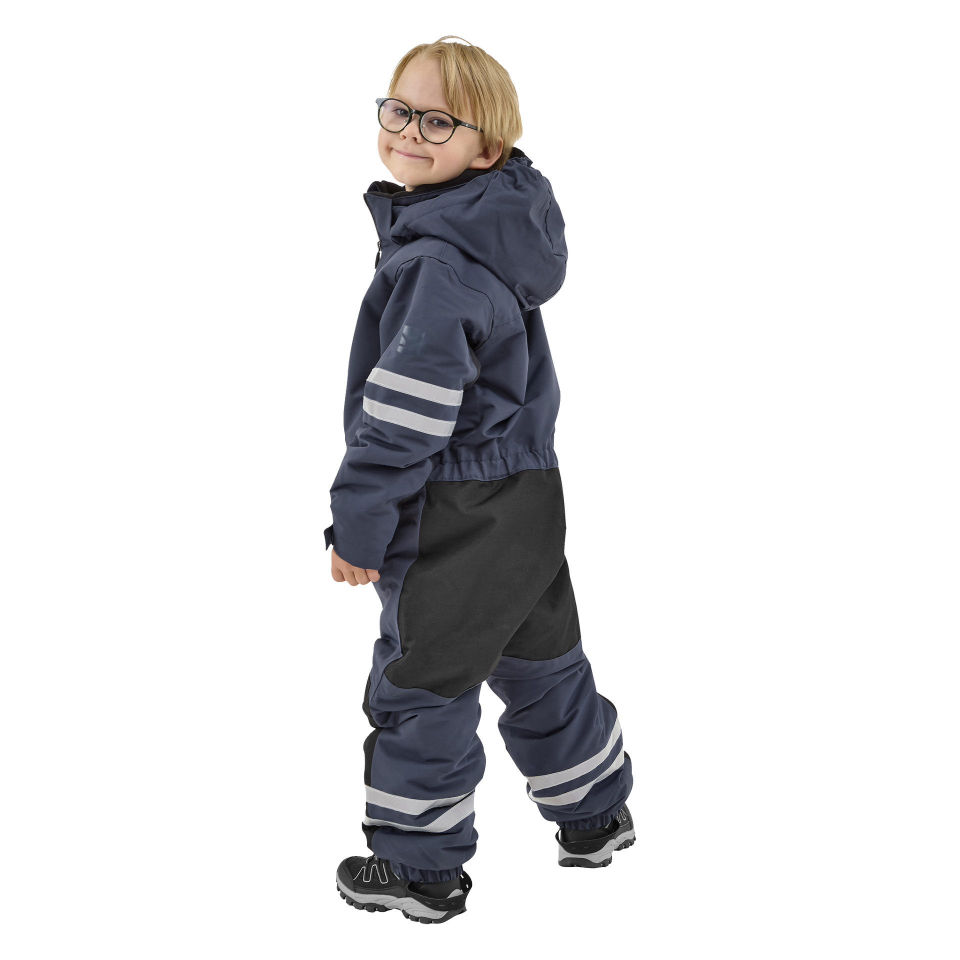 Ett leende ungt barn med blont hår och glasögon, klädd i en marinblå Kayoba Outdoor Dundret-huva och matchande byxor med reflekterande ränder, står lätt på sidan och kastar en blick över axeln.
