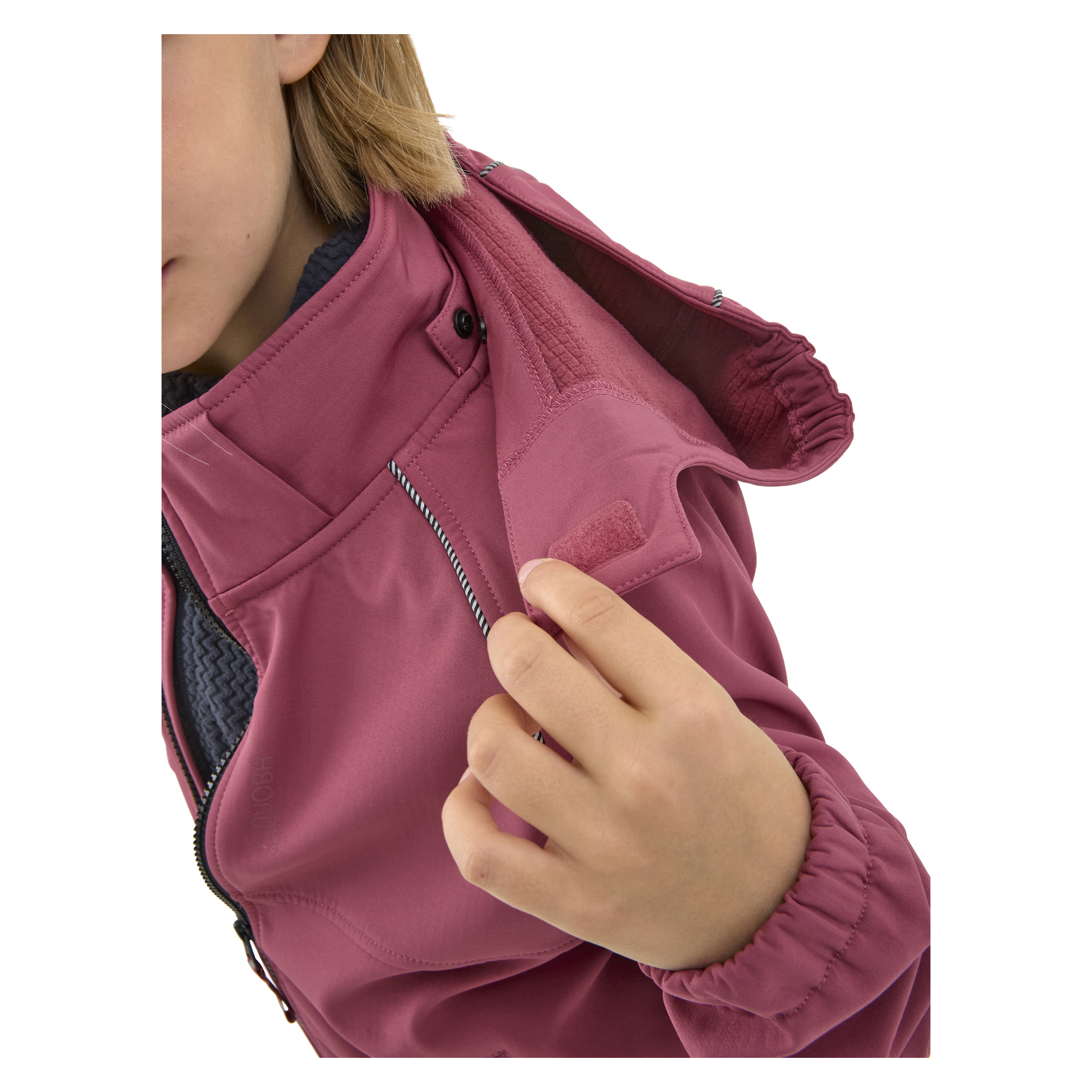 Eine teilweise sichtbare Person öffnet die Velcro-Klappe nahe dem Kragen einer mauve Kayoba Outdoor Kindersoftshell-Softshell-Jacke Kramfors und legt das innere Futter der Jacke frei.
