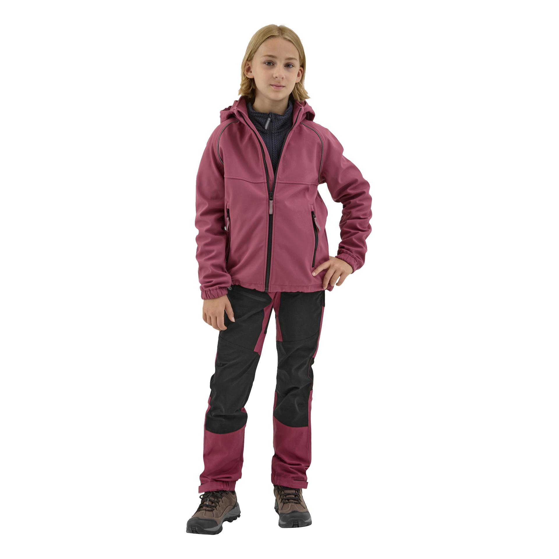 Nuori henkilö, jolla on olkapäille ulottuvat vaaleat hiukset, on pukeutunut Kayoba Outdoor Children's Softshell Jacket Kramforsiin, mustiin ja viininpunaisiin housuihin sekä ruskeisiin vaelluskenkiin, seisoen itsevarmana toinen käsi lanteilla.