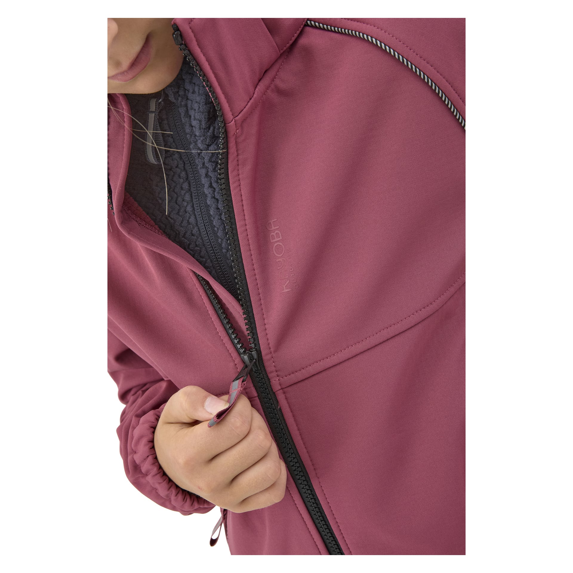 Henkilö, jolla näkyy vain alapuoli kasvot ja käsi, vetää kiinni viininpunaisen Kayoba Outdoor Children's softshell-takin Kramforsin tummanharmaan tekstuurisen kerroksen päälle. Takissa on mustat vetoketjut ja ompeluyksityiskohdat.