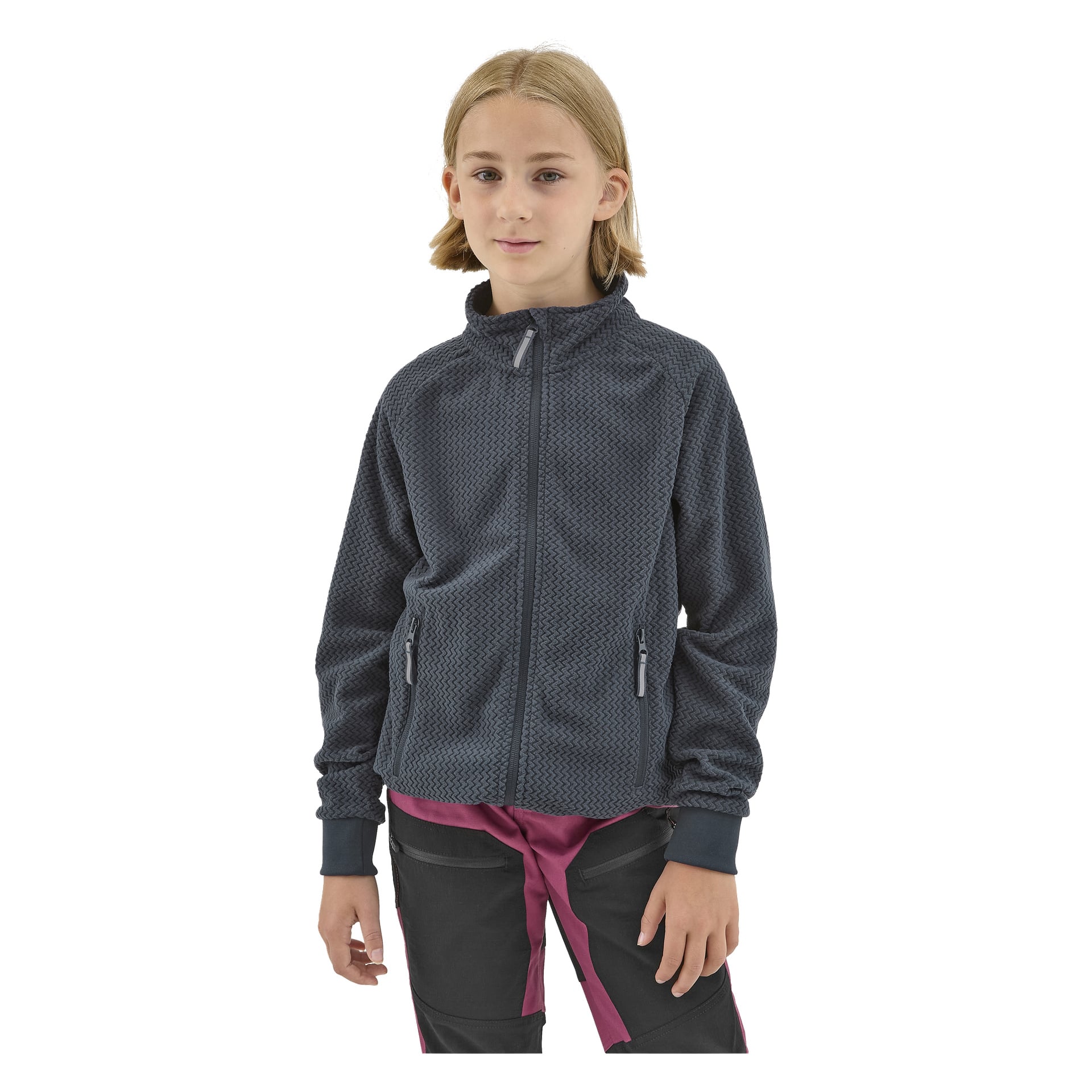 Nuori, jolla on suorat, vaaleanruskeat hiukset, pukeutuu Kayoba Outdoor Children's fleece-takkiin Omberg—tummanharmaaseen, vetoketjulliseen takkiin, jossa on teksturoitu kuvio—yhdistettynä mustiin housuihin, joissa on vaaleanpunaisia yksityiskohtia.