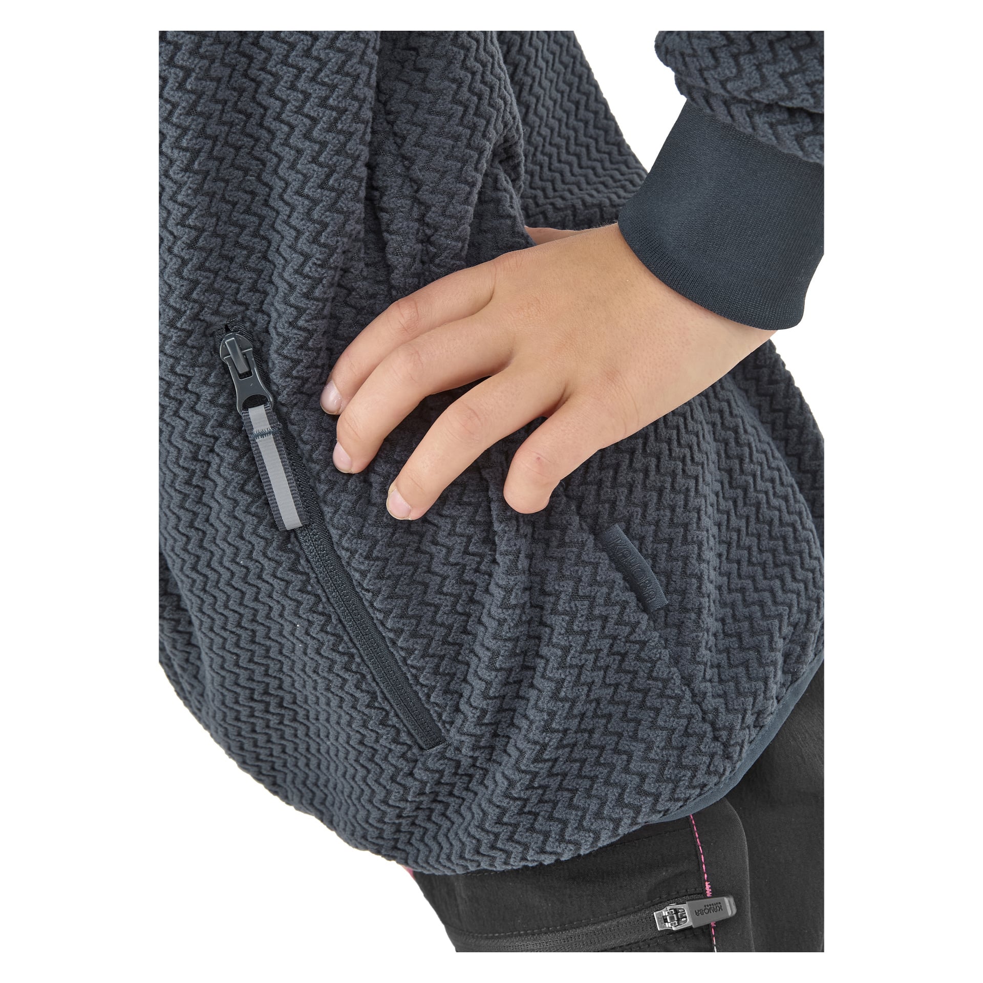 Oikea käsi asetetaan Kayoba Outdoor Children's fleece -takki Ombergin sivulle, jossa on teksturoitu tumma chevron-kuvio ja näkyvä etutasku. Mustien housujen osasto näkyy myös.