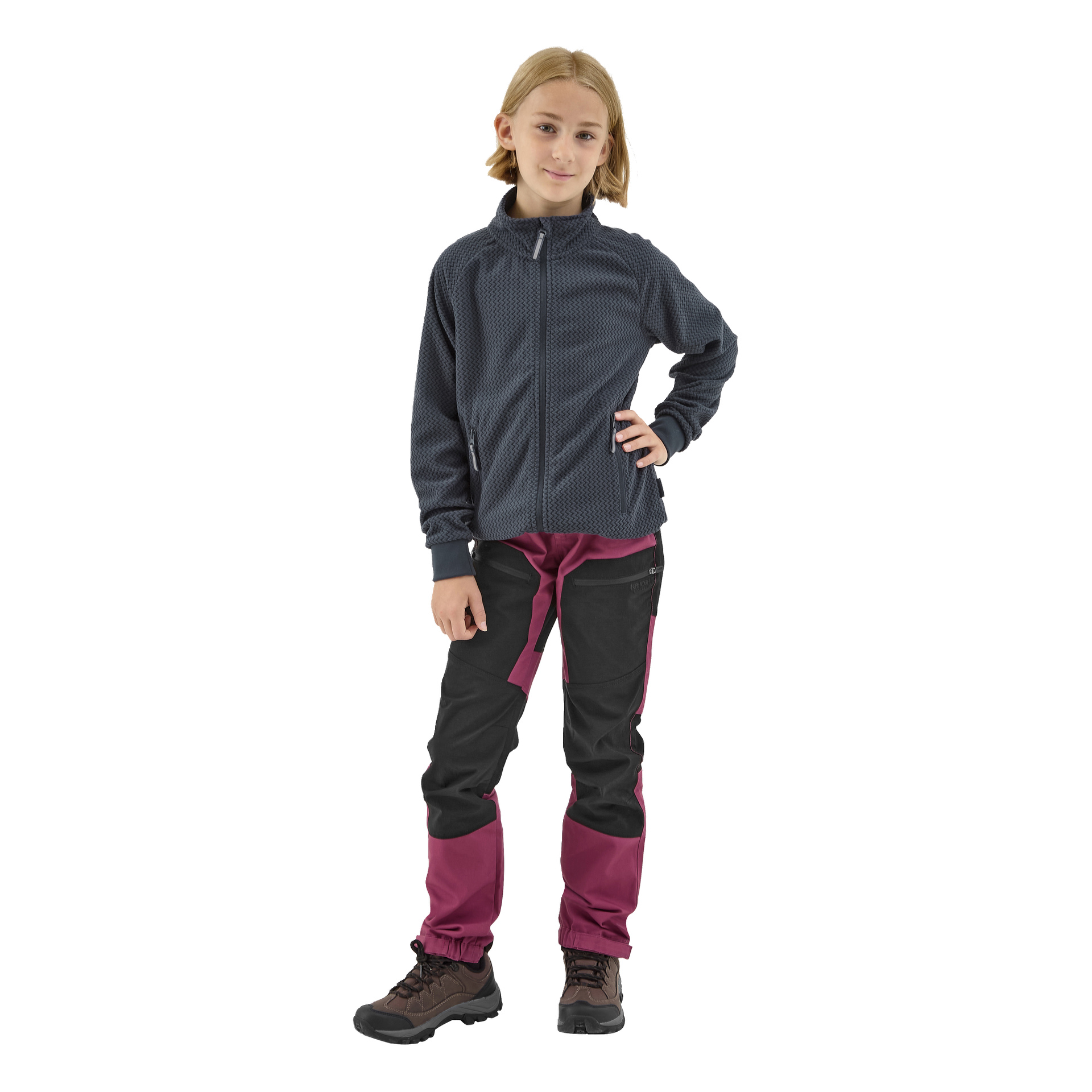 Lyhyt vaaleahiuksinen lapsi on pukeutunut Kayoba Outdoor Children's fleece -takkiin Omberg, yhdistettynä mustiin ja magentanvärisiin ulkohousuihin sekä ruskeisiin vaelluskenkiin. Lapsi seisoo itsevarmasti, toinen käsi lanteella, kameraa kohti.