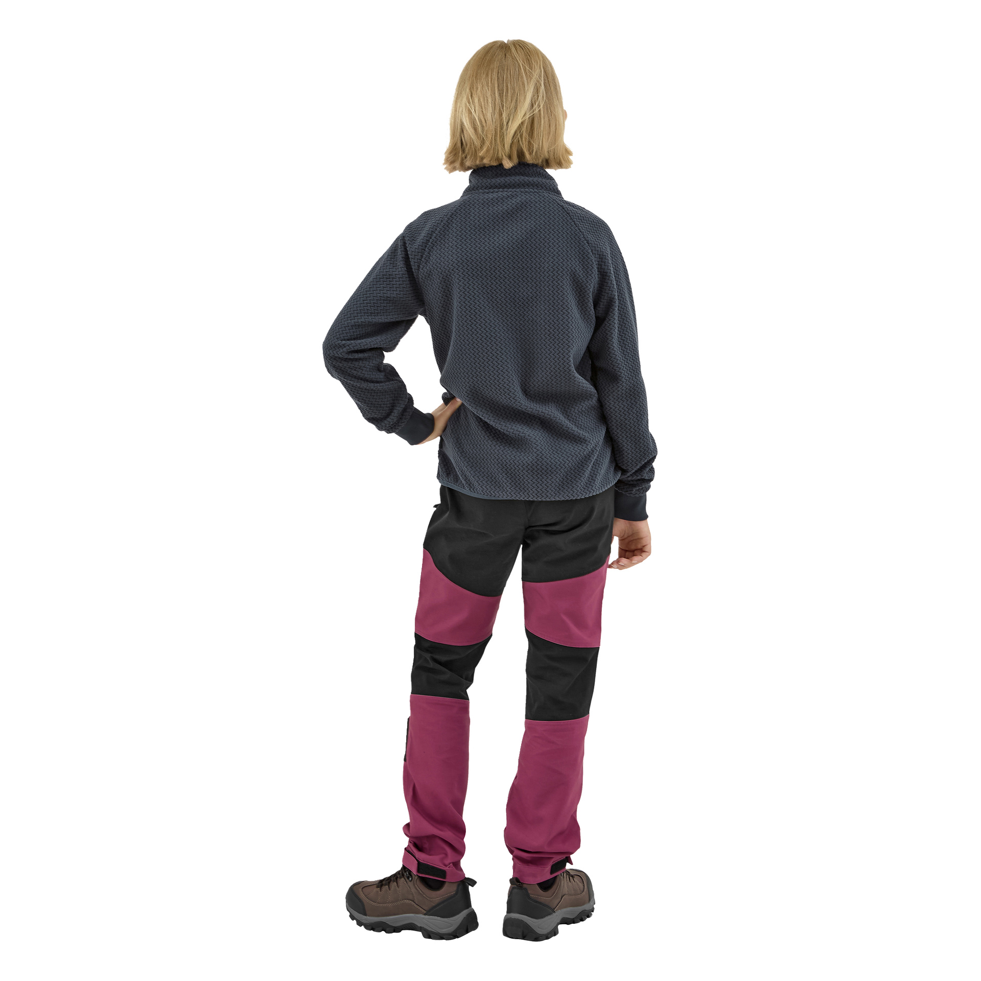 Vaaleahiuksinen lapsi nähdään takaapäin, pukeutuneena Kayoba Outdoor Children's fleece -takkiin Omberg, mustat ja magentan väriset housut ja ruskeat vaelluskengät. Vasen käsi lepää lanteella ja oikea käsi roikkuu löysästi sivulla.