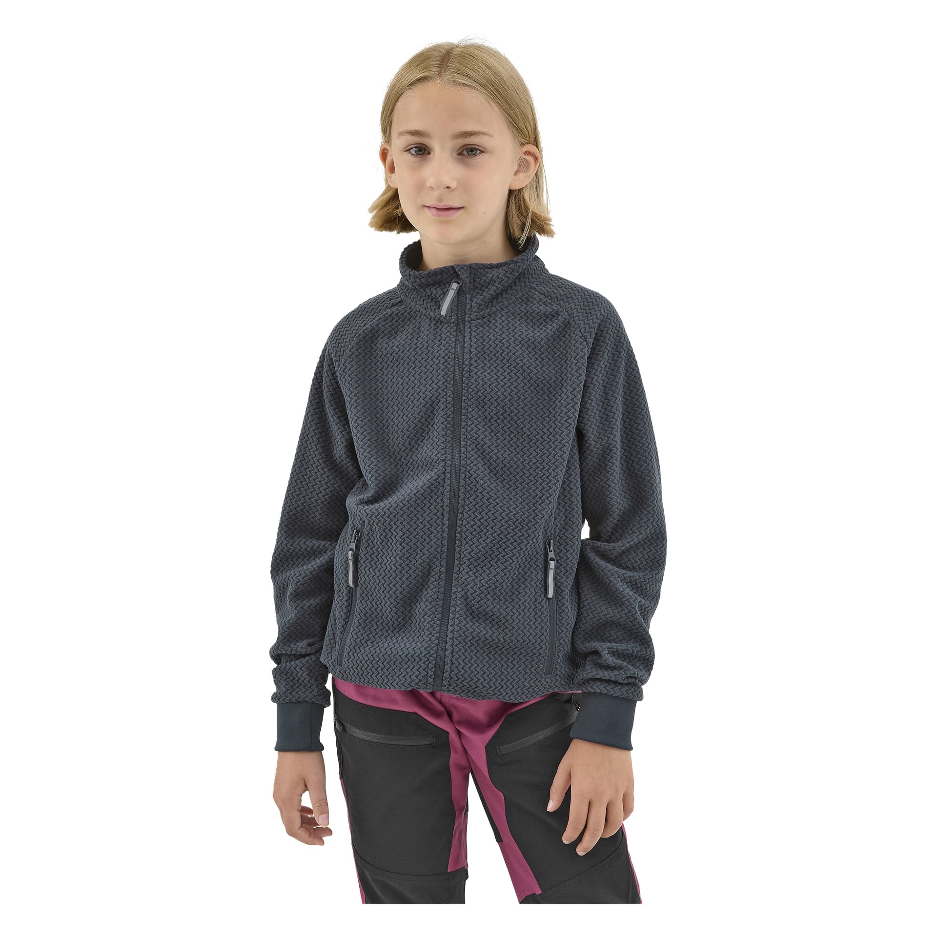 Nuori, jolla on olkapäille ulottuvat vaaleat hiukset, käyttää Kayoba Outdoor Children's fleece-takkia Omberg, tumman tekstuurisen vetoketjun päällä, kerrostettuna viininpunaisen paidan ja mustien housujen päälle, joissa on viininpunaisia yksityiskohtia.