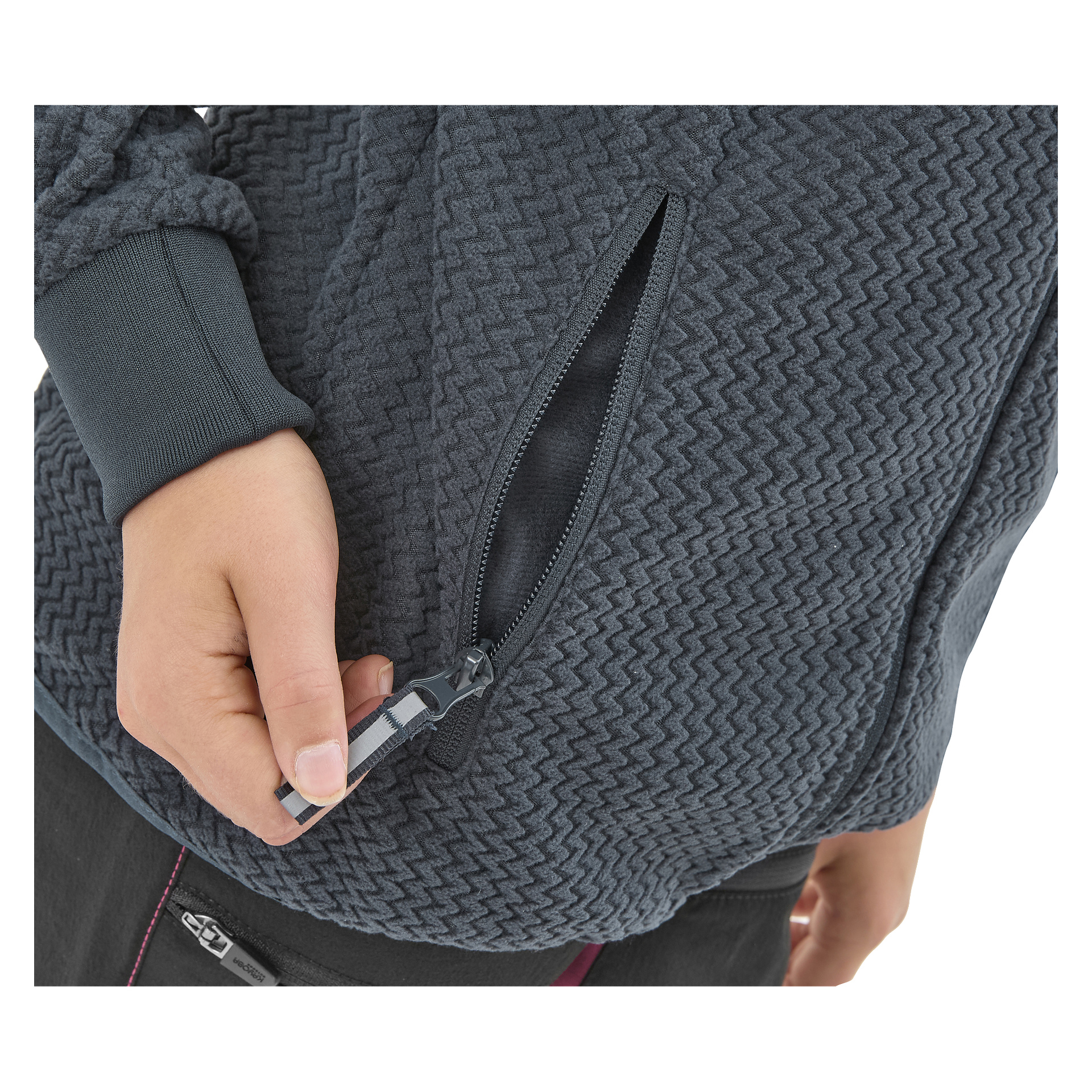 Joku avaa Kayoba Outdoor Children's fleece-takin Ombergin sivutaskua, joka on tummanharmaa tekstuuri. Kuvassa näkyy vain heidän kätensä, osa takista ja osa housuista.
