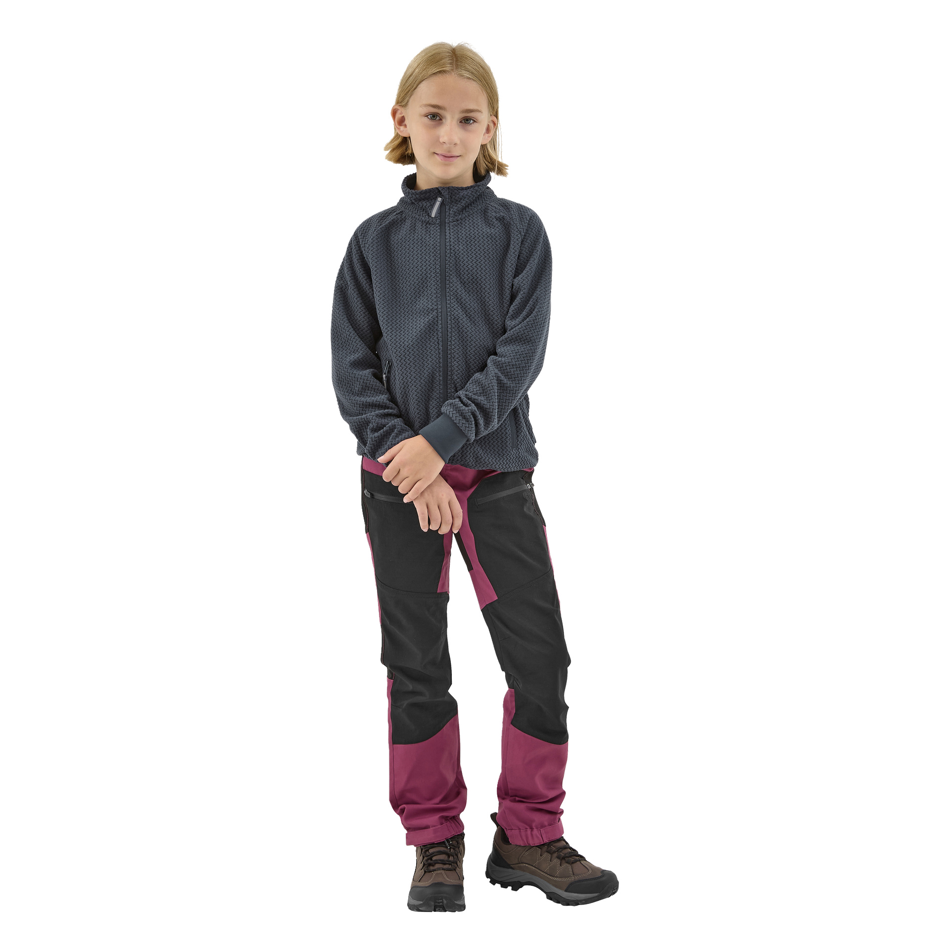 Vaaleanruskeatukkainen lapsi seisoo kasvot eteenpäin, pukeutuneena Kayoba Outdoor Children's fleece -takki Ombergiin, mustiin ja magentanvärisiin housuihin sekä ruskeisiin vaelluskengiin. Lapsen kädet ovat varovasti ristissä edessä.