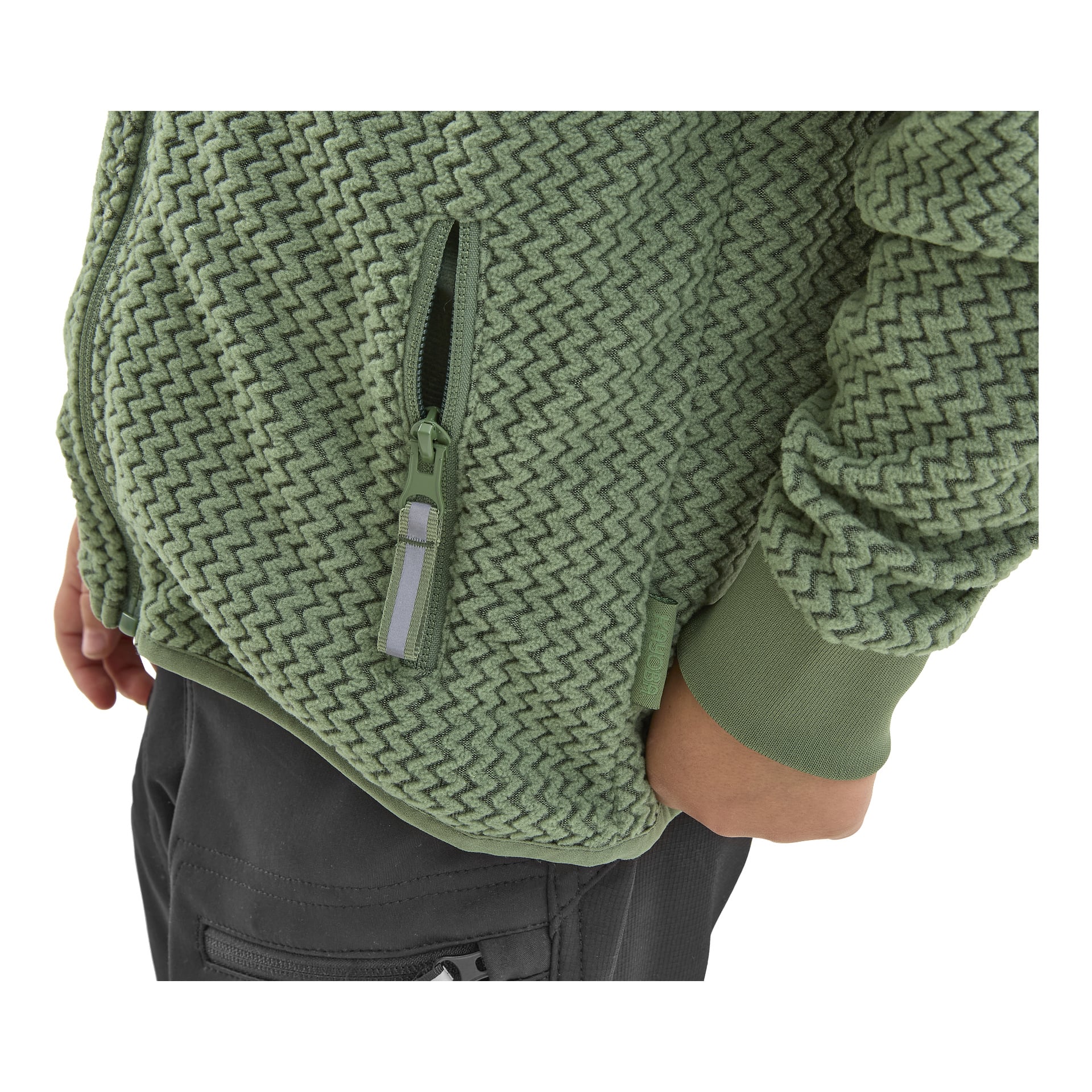 Lapsi pukeutuneena vihreään Kayoba Outdoor Children's Omberg -fleecetakkiin ja tummiin housuihin, joissa on sivutasku, näkyvissä alavartalosta alaspäin. Painopiste on takin teksturoidussa fleece-materiaalissa ja näkyvissä vetoketjun piirteissä.
