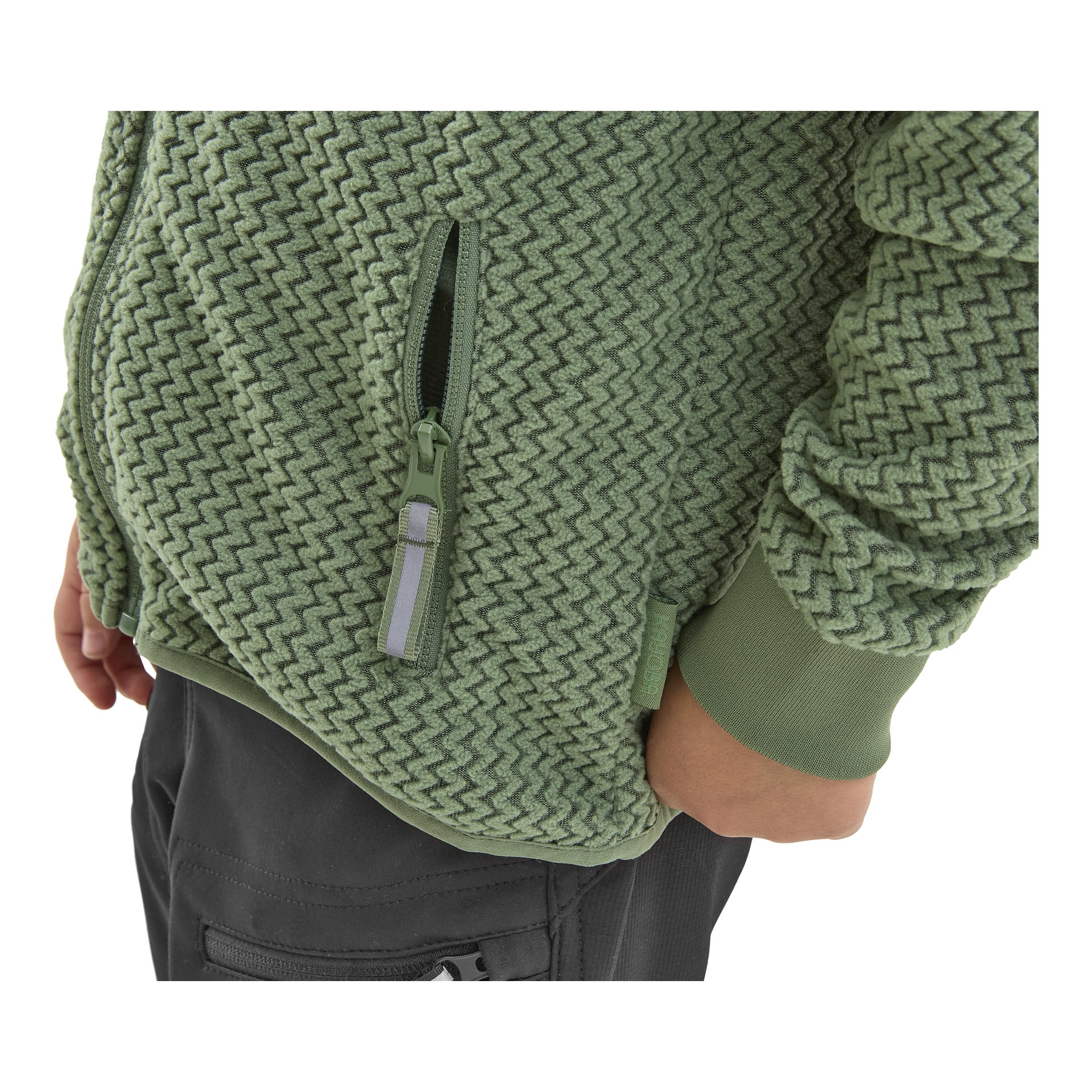 Lapsi pukeutuneena harmaisiin housuihin ja Kayoba Outdoor Omberg -fleecetakkiin, jossa on teksturoitu vihreä siksak-kuvio. Takin vetoketjullinen tasku on osittain auki. Kuvassa näkyy vain lapsen vartalo ja osa käsivarsista, toinen käsi rentoutuneena sivuilla.