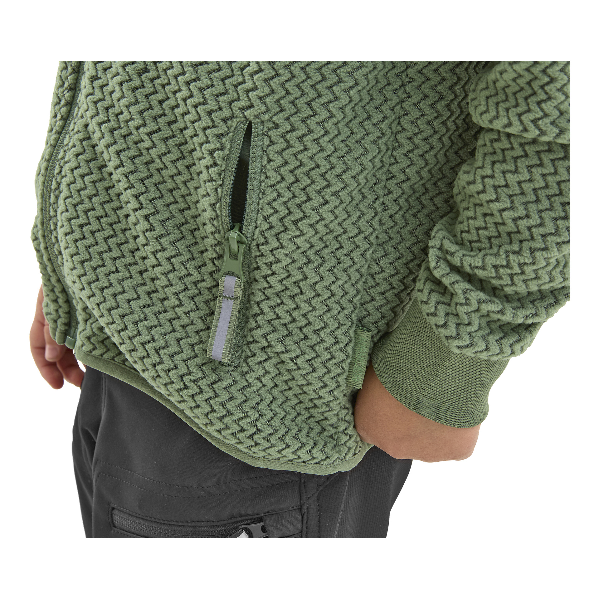 Lapsi mallintaa Kayoba Outdoor Children's Omberg -fleecetakkia, seisoo toinen käsi taskussa. Kuvassa näkyy vihreä teksturoitu vetoketjullinen takki ja tummat housut, mikä kiinnittää huomion vetoketjulliseen taskuun ja hihan sekä vartalon uurteisiin kalvosimiin. Painopiste on lapsen keskivartalossa ja käsivarressa.