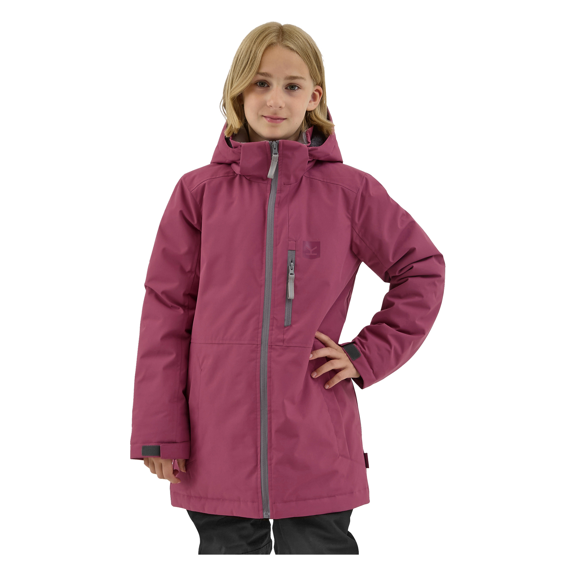 En ung person tittar framåt med ena handen vilande på höften, klädd i en vinröd Kayoba Outdoor Children's vinterjacka Kläppen som har huva, dragkedja fram och bröstficka med dragkedja.