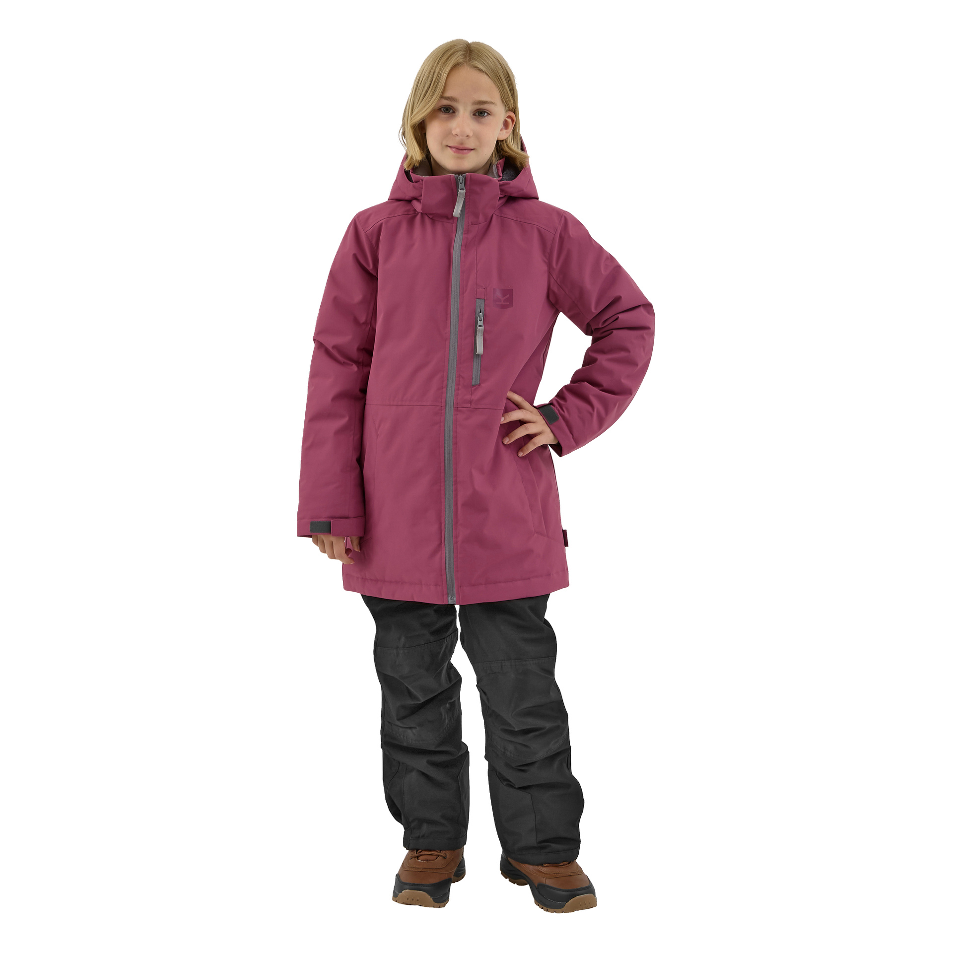 En ung person med långt blont hår står vänd framåt, klädd i en magenta Kayoba Outdoor Children's vinterjacka Kläppen, svarta byxor och bruna stövlar. En hand placeras på höften.