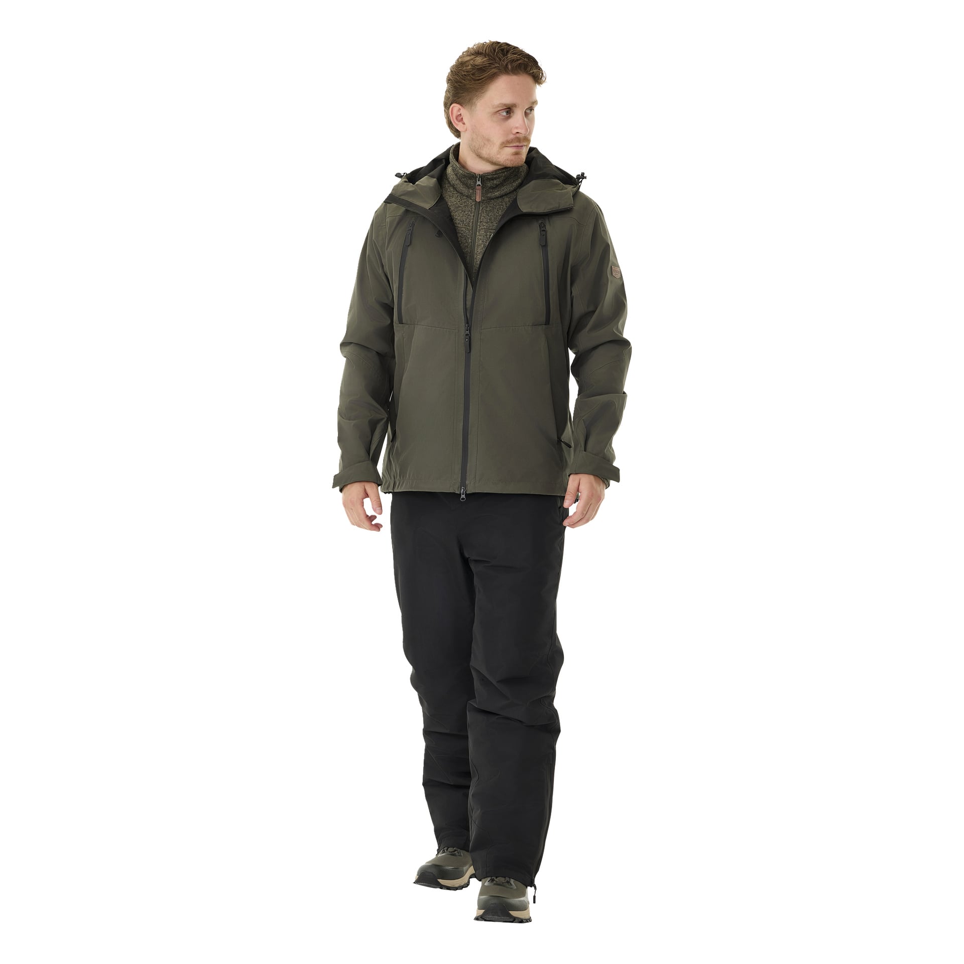 Mies, jolla on lyhyet, vaaleanruskeat hiukset ja neutraali ulkonäkö, seisoo hieman sivulle, pukeutuneena Kiruna 2.0 -pehmustettuihin miesten housuihin Kayoba Outdoorilta, vihreään hupparitakkiin, oliivinväriseen neuleeseen ja mustiin lenkkareihin.