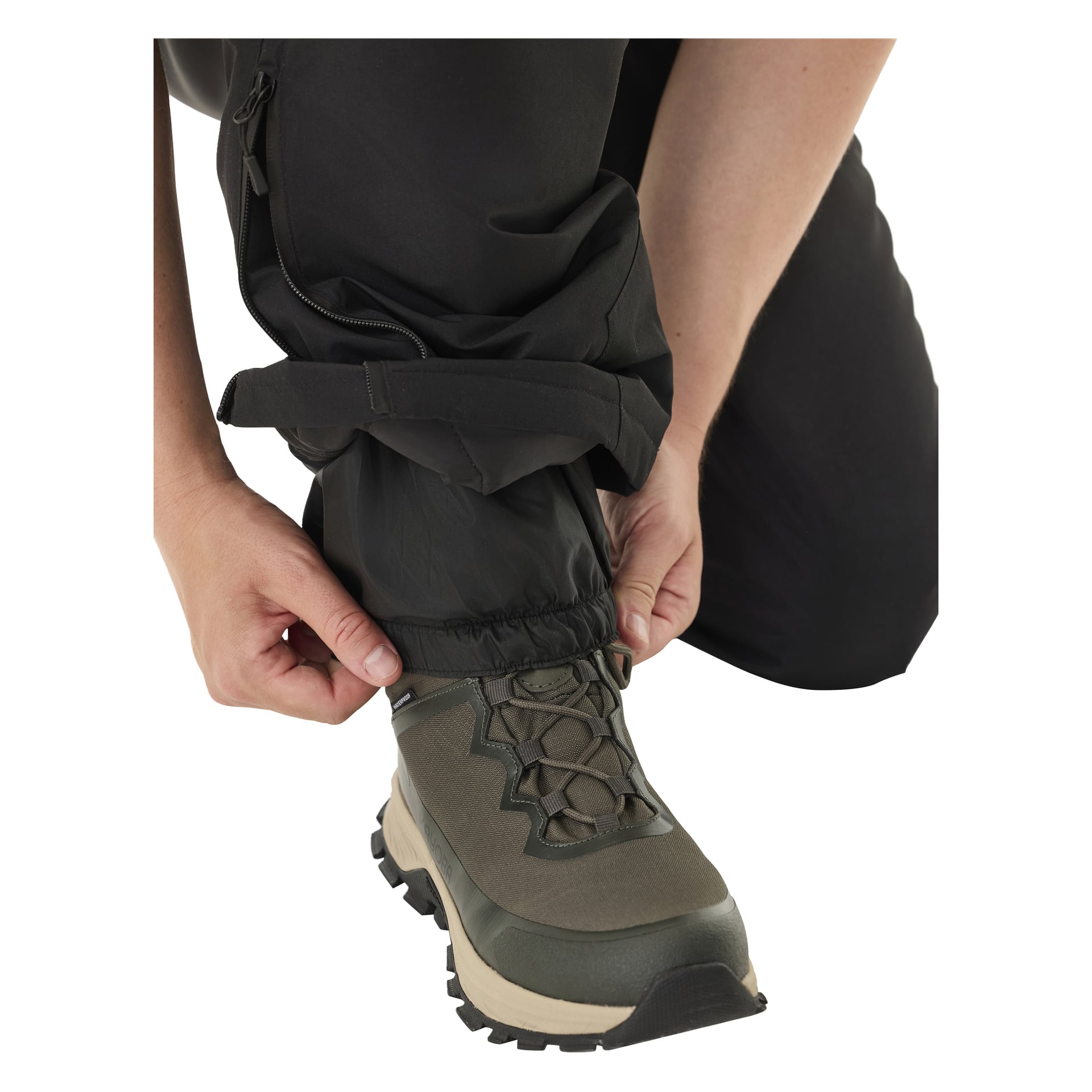 Henkilö polvistuu, vetäen Kayoba Outdoor Padded Kiruna 2.0 -miesten housujensa kuminauhan oliivinvihreän vaelluskengän päälle, jossa on ruskeat ja mustat yksityiskohdat. Housujen vedenkestävä materiaali näkyy selvästi.