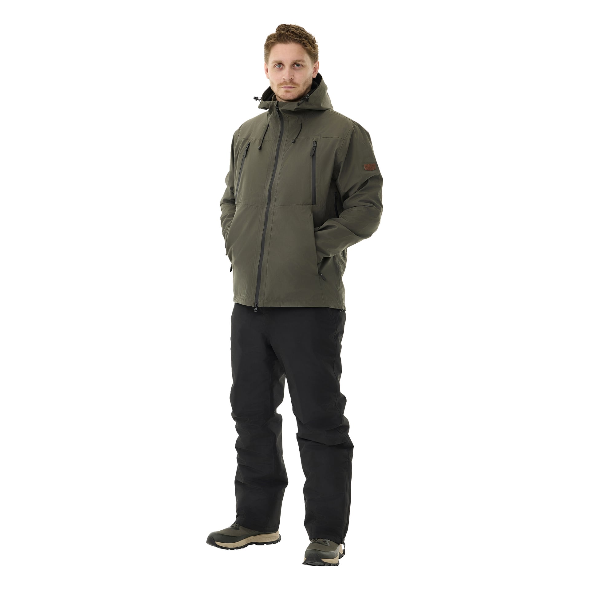 Mies, jolla on neutraali ilme, seisoo eteenpäin, kädet taskuissa. Hänellä on mustat Kayoba Outdoor -pehmussuojahousut miehille Kiruna 2.0, oliivinvihreä hupparitakki sekä beiget ja mustat kengät.