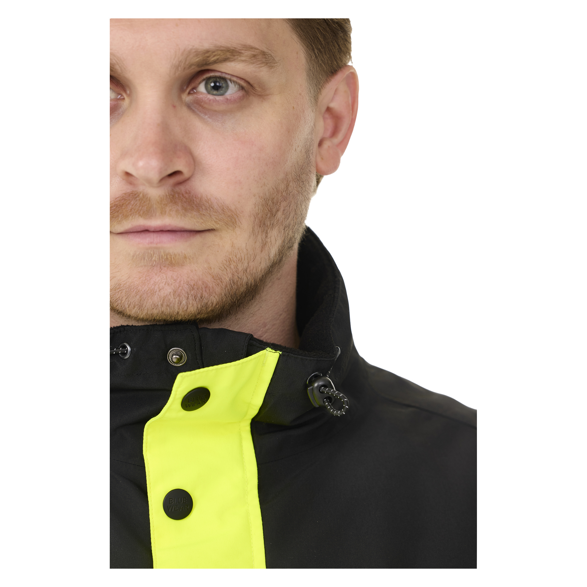 Lähikuva miehen kasvoista ja ylävartalosta. Hänellä on vaaleat kasvojen karvat, siniset silmät ja hänellä on päällään Bluewear Pehmustettu hi-vis-takki Lawson, jossa on kirkkaan keltainen kaulusyksityiskohta.