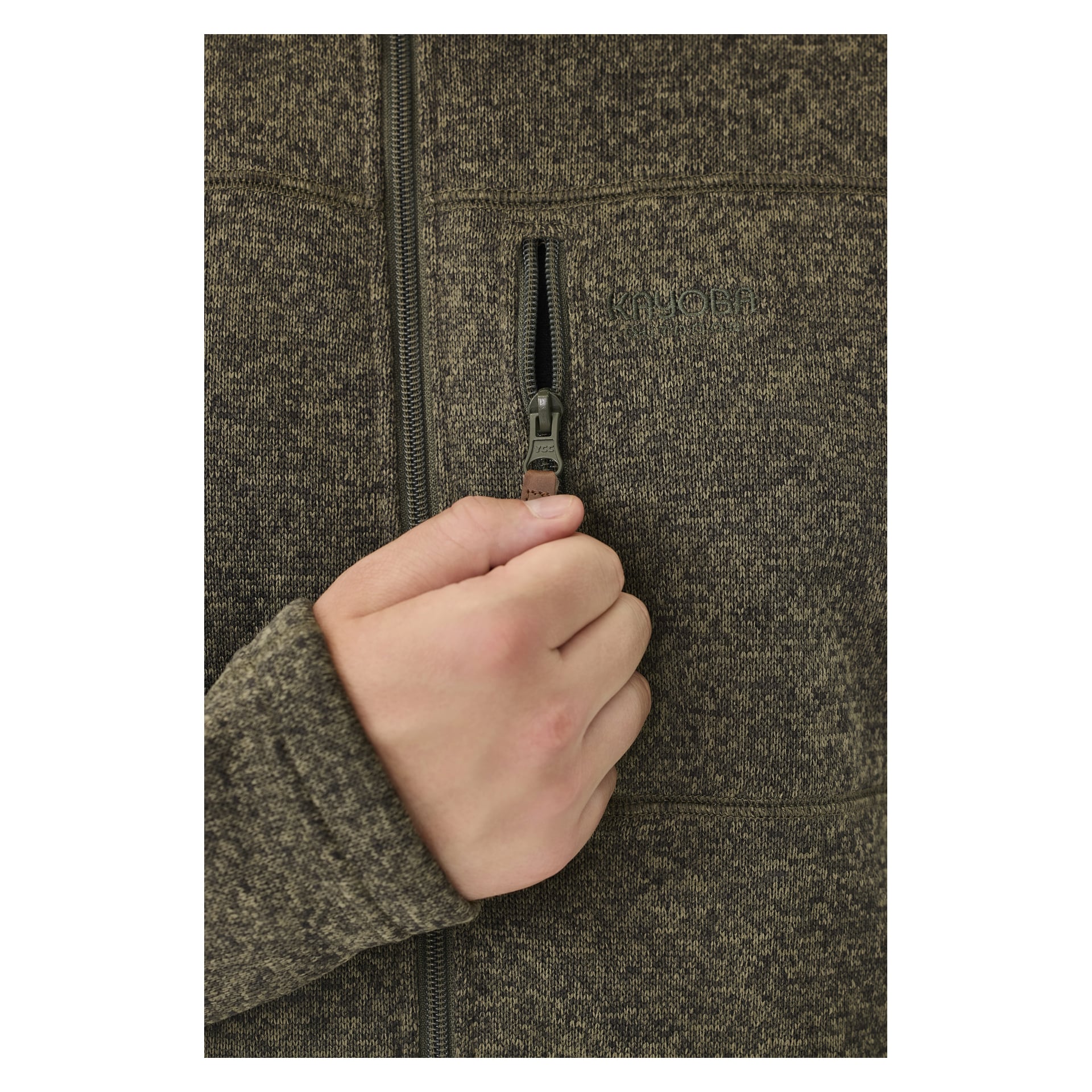 Et nærbilde viser en hånd som holder glidelåsen på en Kayoba Outdoor Knitted fleecejakke for menn, Malå i flekkete brun, delvis åpnet en liten vertikal brystlomme. Den detaljerte strikketeksturen og stoffveven er tydelig synlig.