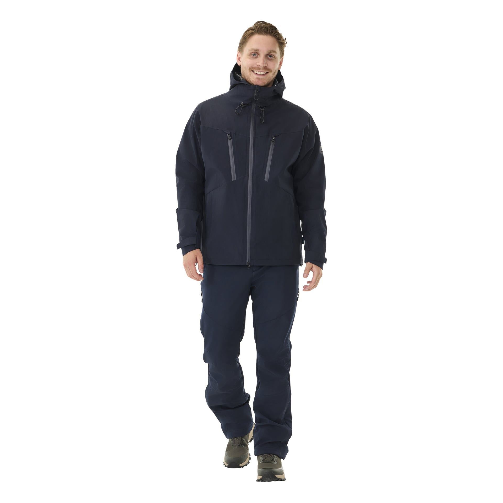 En person står med blicken framåt, klädd i Kayoba Outdoor Men's Sarek shelljacka i mörkt marinblått, med huva, synliga dragkedjor och fickor. Outfiten kompletteras med matchande marinblå byxor och grå skor.