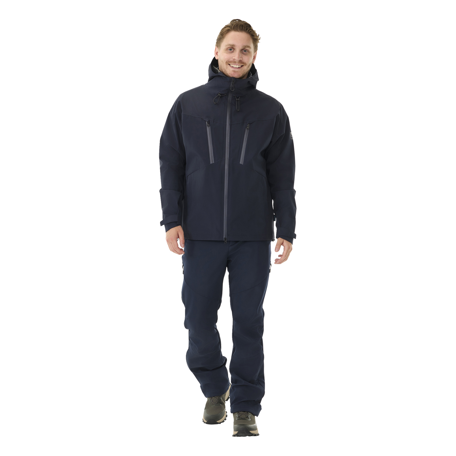 En mann vender fremover i Kayoba Outdoor Men's Sarek shelljakke i mørk marineblå, kombinert med matchende marineblå bukser og svarte tursko. Han smiler med hendene avslappet langs sidene.