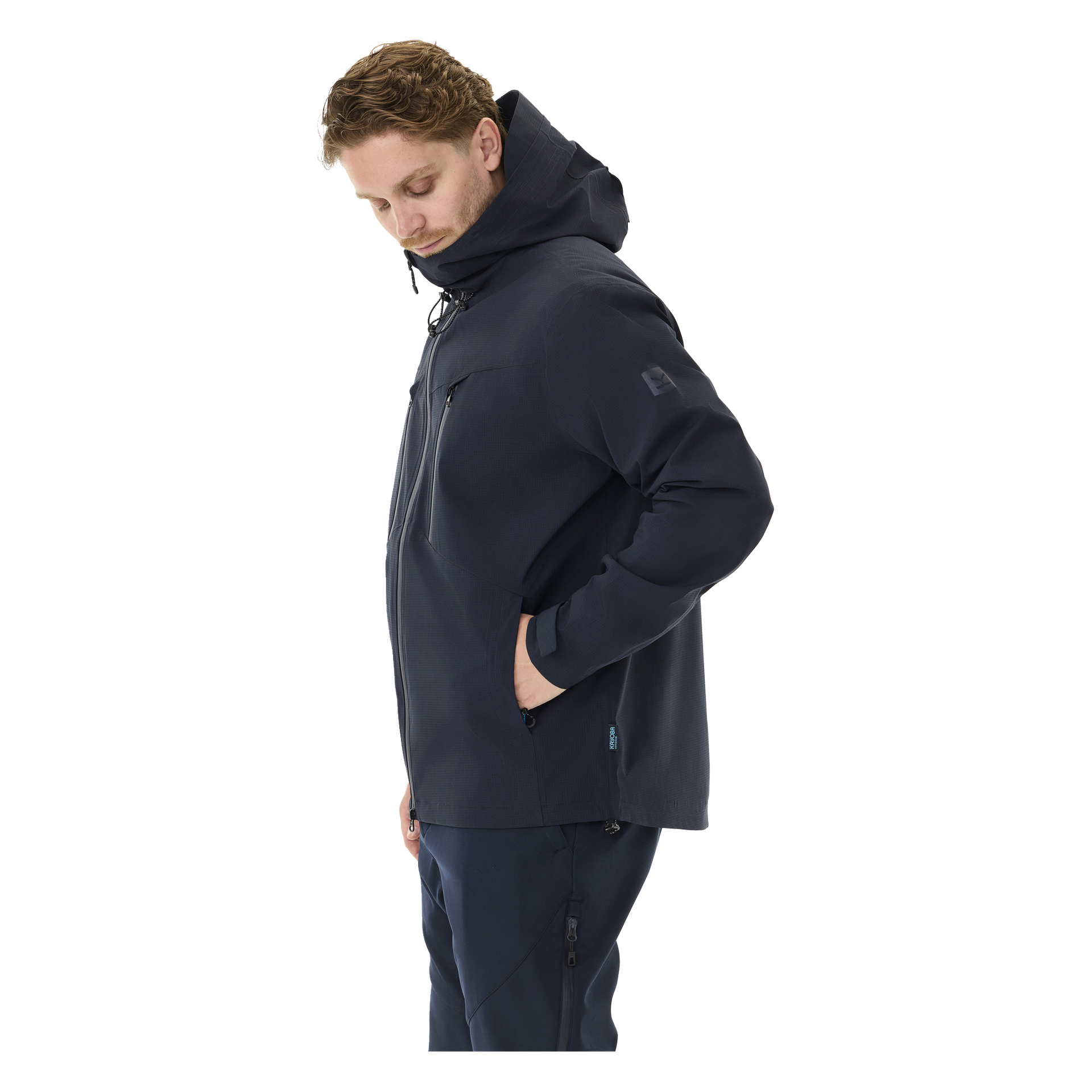 En person står i profil, kledd i Kayoba Outdoor Men's shell-jakke, Sarek i mørk marineblå og matchende bukser, en hånd stukket i lommen og blikket rettet nedover.