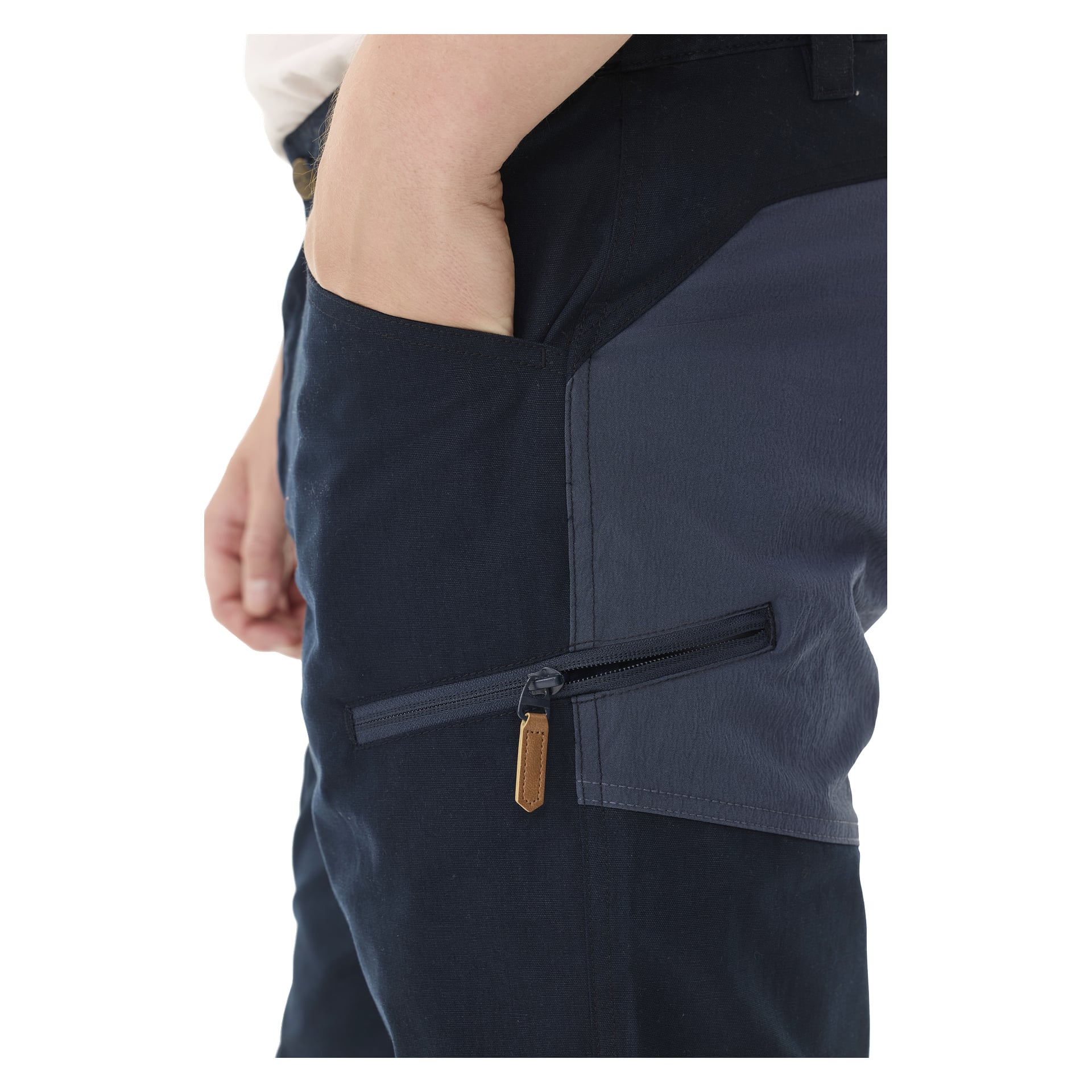 Eine Person trägt die Kayoba Outdoor Men Älvdalen 2.0 Hosen, dunkelblau mit grauem Seitenteil und einer horizontalen Reißverschlusstasche mit einem braunen Zugtab. Eine Hand steckt in die Tasche.