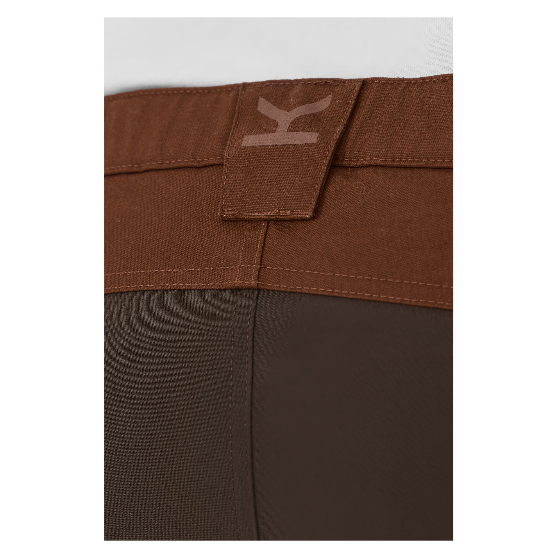 Detaljert bilde av linningen på Men's Älvdalen 2.0 Outdoor Trousers fra Kayoba Outdoor, med en stoffløkke og en karakteristisk K-logo nær sømmen. Buksene er laget av to kontrasterende brune stoffer.