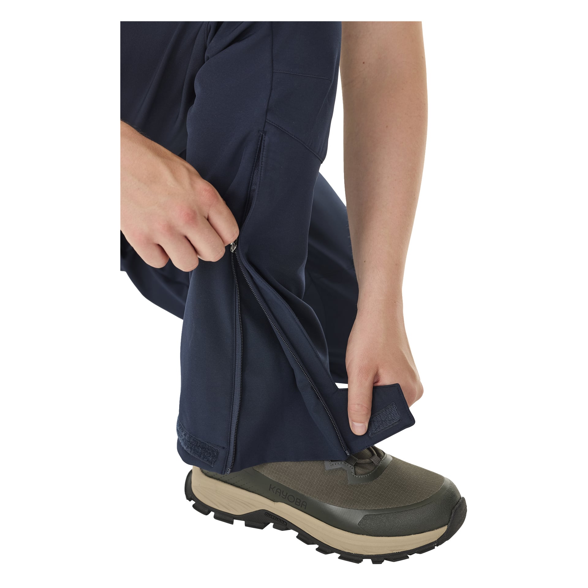 Henkilö tummansinisissä Kayoba Outdoor Men's -vuorisissa pehmeäkuorisissa housuissa kyykistyy ja avaa toisen housunlahkeen vetoketjun paljastaakseen vihreät vaelluskengät, käsi vetoketjulla.