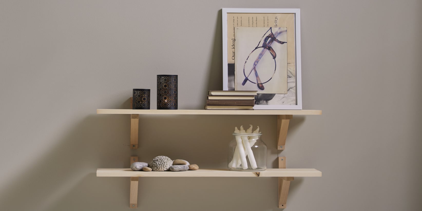 Två ljusa trähyllor monterade på en beige vägg visar ljus, böcker, polerade stenar och en inramad bild med glasögon. Plaggen är prydligt arrangerade för ett minimalistiskt och organiserat utseende.