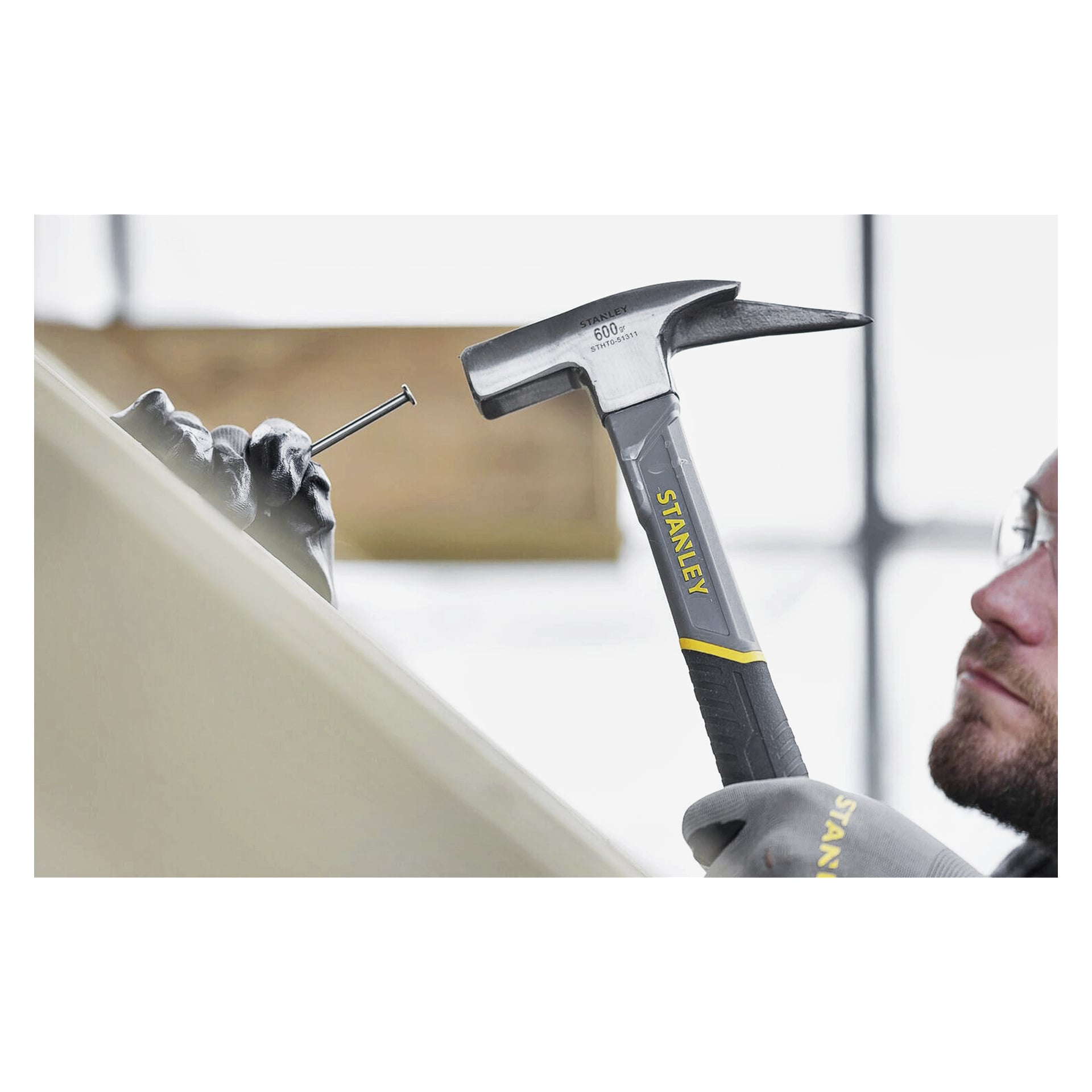 Eine behandschuhte Person hält mit einer Hand einen Nagel auf einer Holzoberfläche, während sie sich darauf vorbereitet, mit dem Stanley Roofing Hammer STHT0-51311 in der anderen Hand darauf zu schlagen. Der Hintergrund wirkt unscharf.
