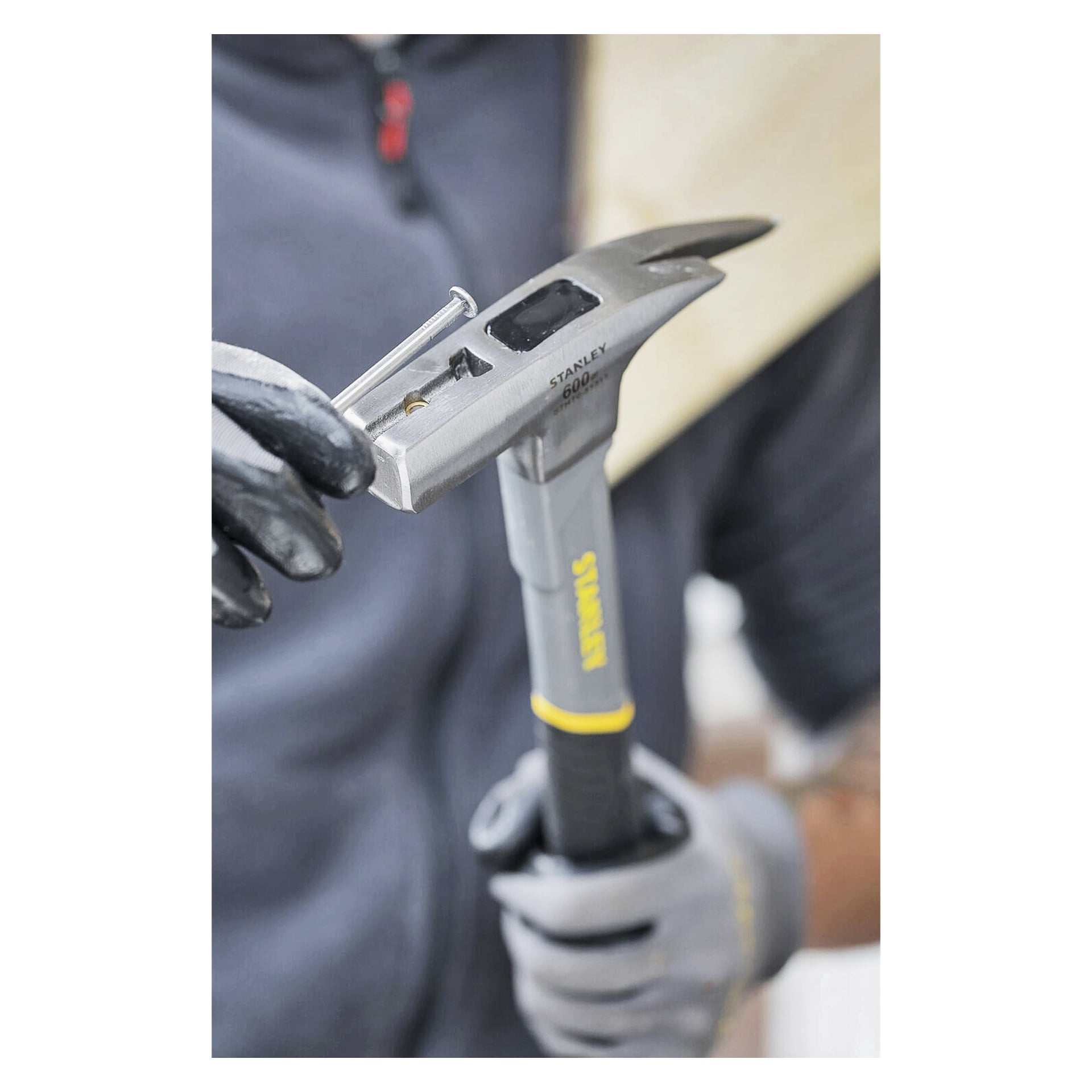 Eine Person mit Handschuhen und einer dunklen Reißverschlussjacke benutzt das Klauenende eines Stanley Roofing Hammer STHT0-51311, um einen Nagel aus dem Holz zu ziehen, wobei sie sich auf ihre Hände und den Hammer konzentriert.