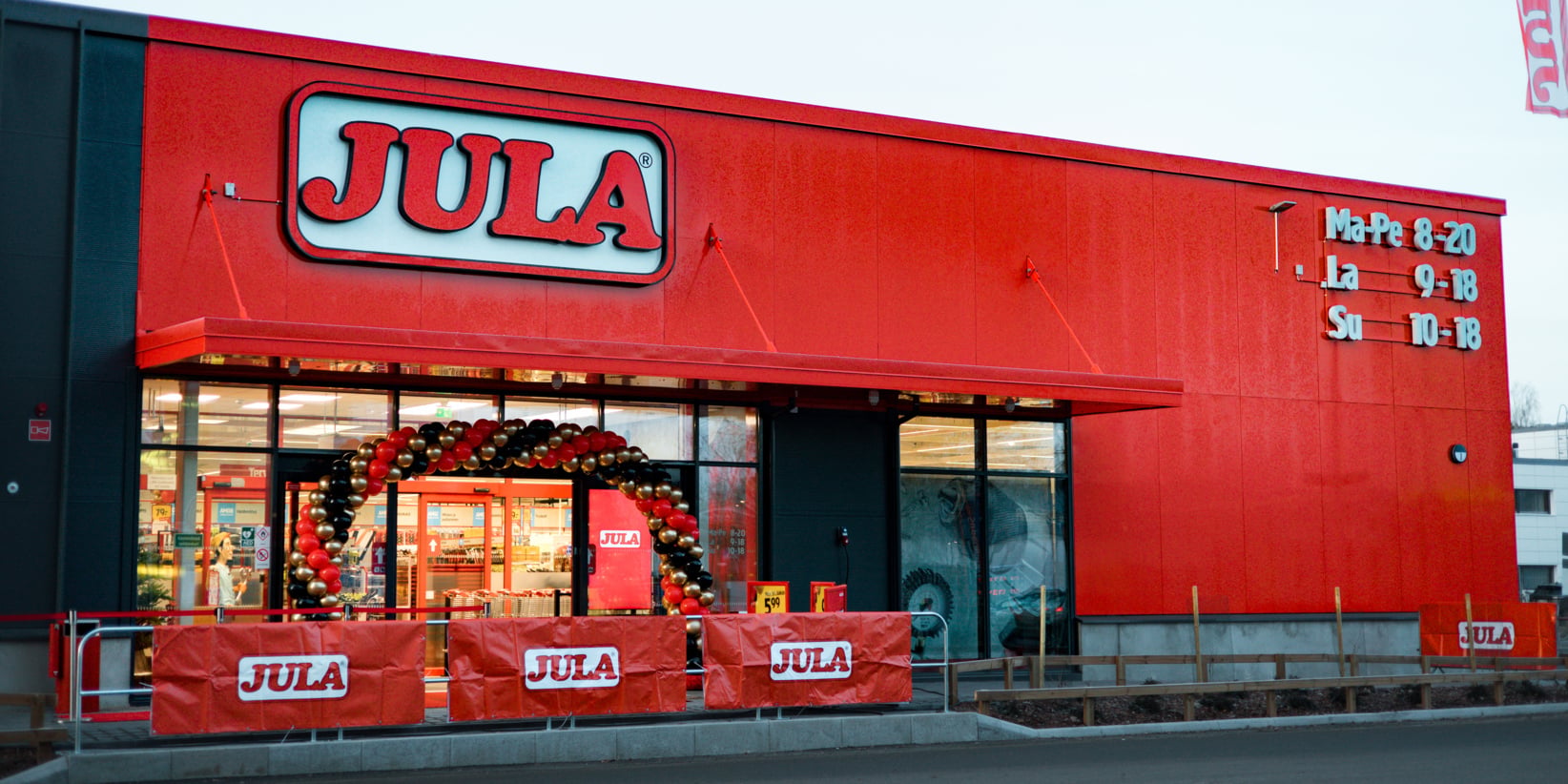 Punainen Jula-kauppa, jossa on näkyvät logokyltit. Punaiset, mustat ja valkoiset ilmapallokoristeet kehystävät sisäänkäyntiä. Aukioloajat on merkitty oven vieressä olevassa seinässä.