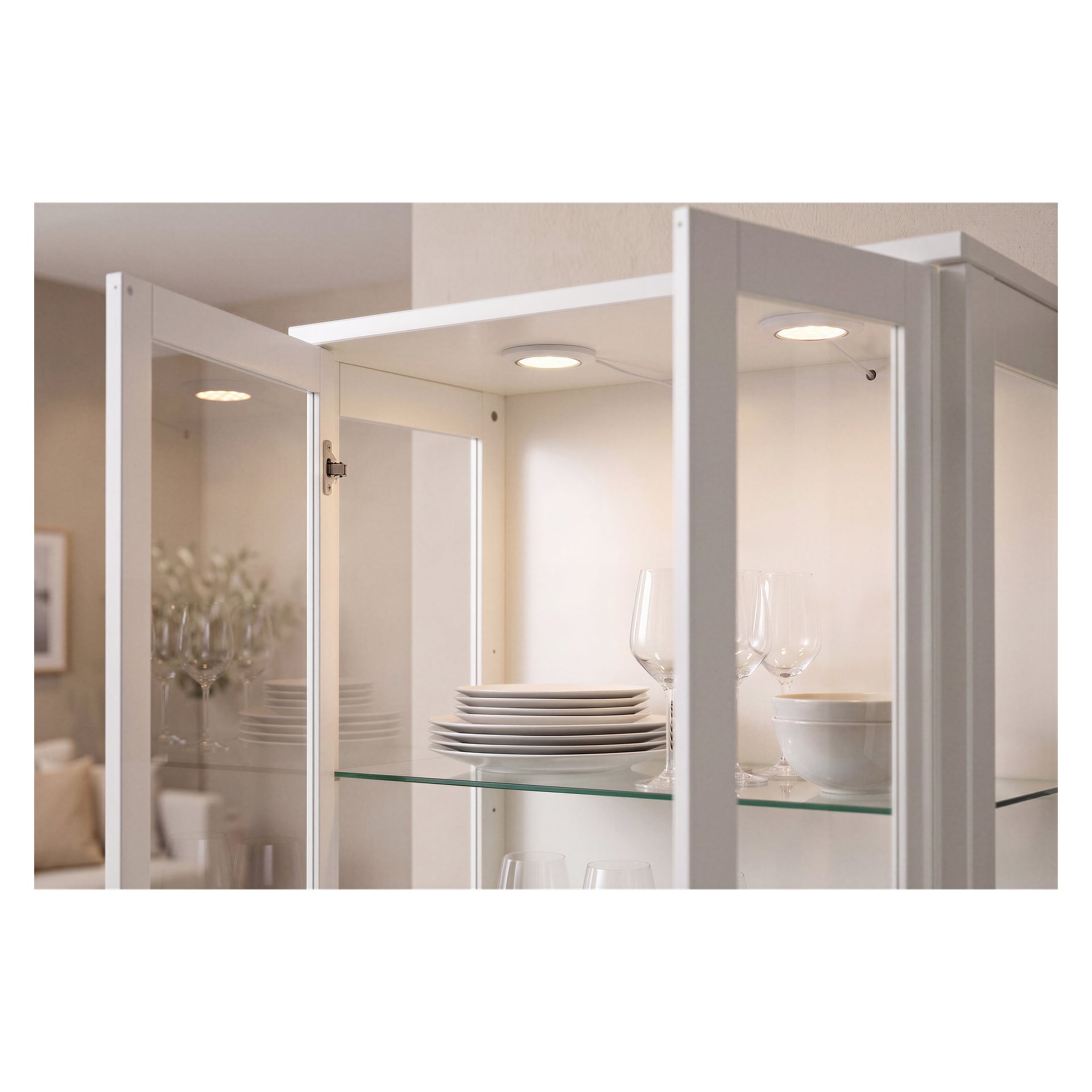 Et glassskap med åpne dører viser pent arrangerte hvite tallerkener, boller og vinglass på de klare hyllene. Over dem kaster en Anslut Downlight LED 3-pakning lys over gjenstandene. Et mykt uklart oppholdsrom vises i bakgrunnen.