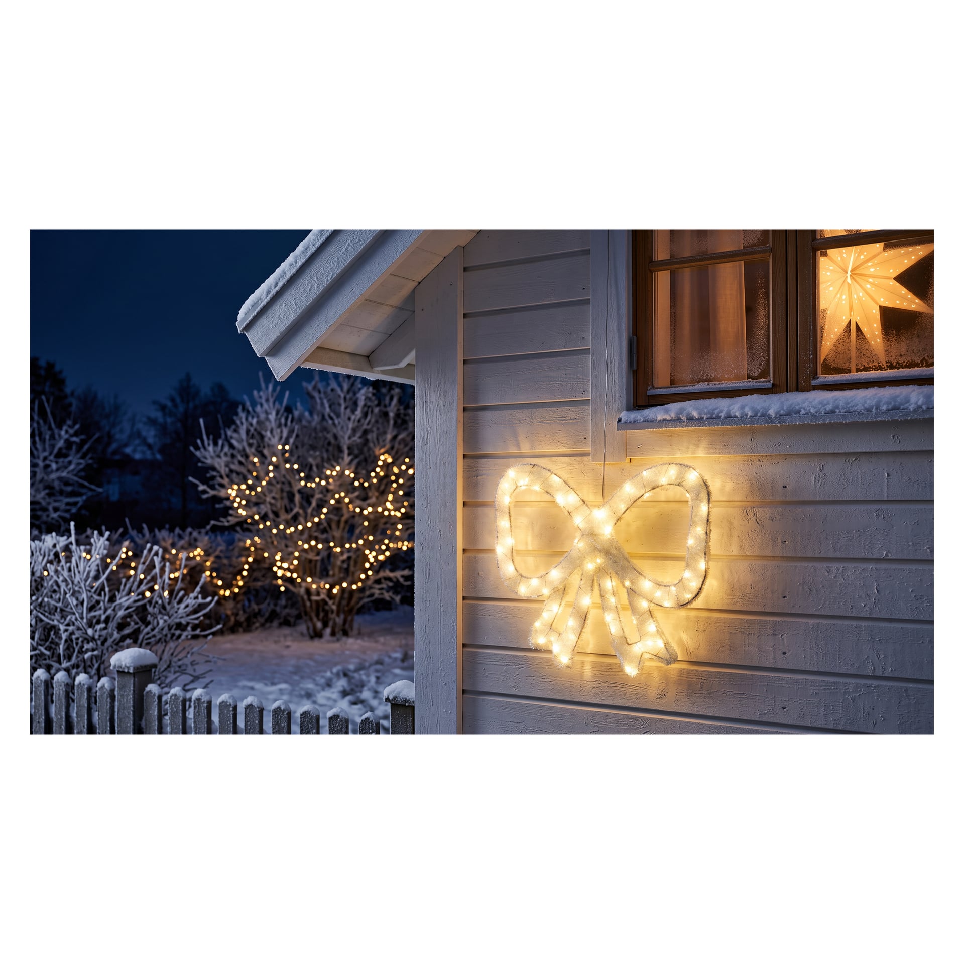 Ett vitt hus med snö på taket och fönsterbrädan visar en EKVIP-julrosett upplyst med LED-lampor på väggen. I bakgrunden står träd dekorerade med ljusslingor bland snötäckt mark och grenar.
