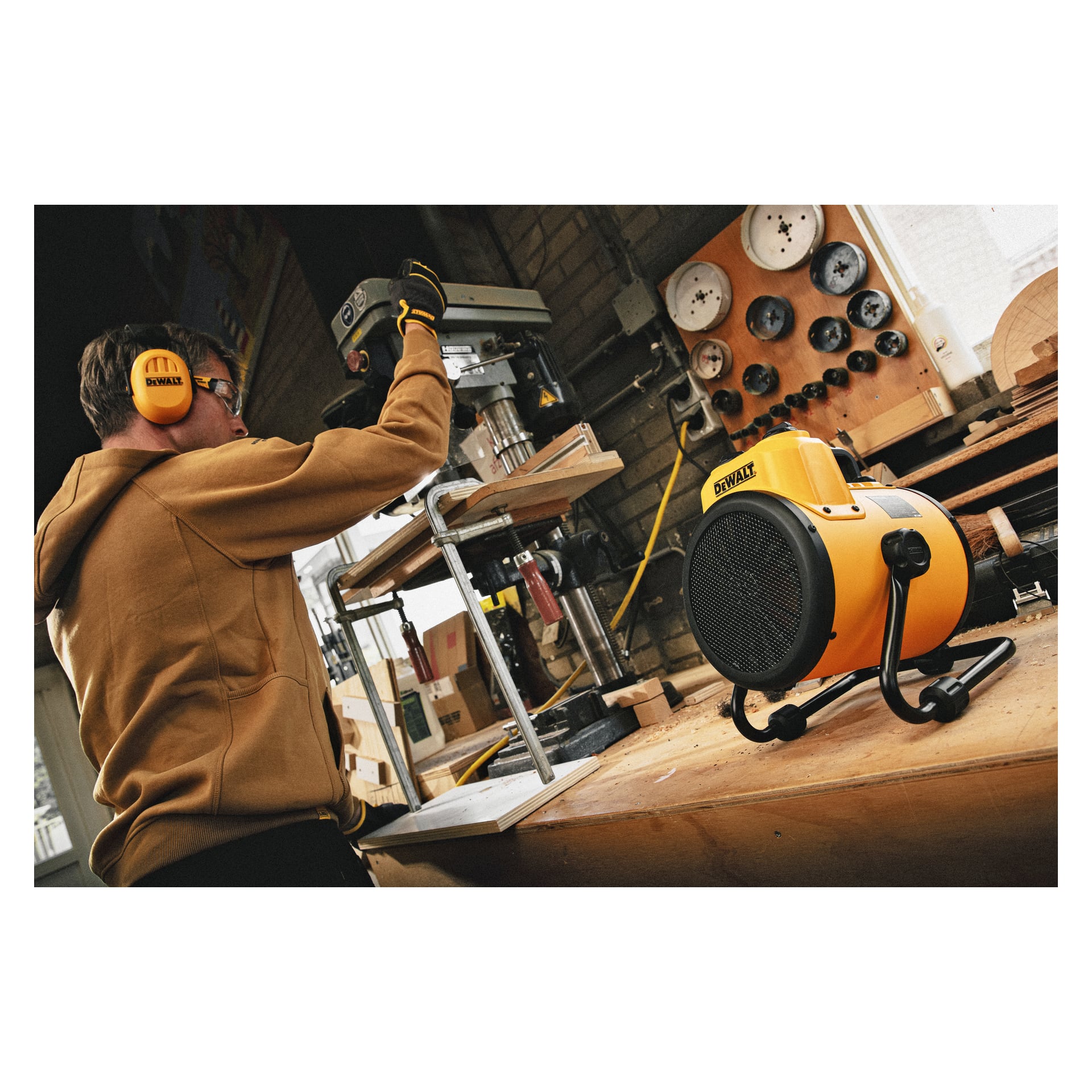 En person med öronskydd och handskar använder en pelarborrmaskin i en verkstad. På träarbetsbänken framför finns en gul DeWalt Heater Fan DXJHXA0002E. Bakom dem syns diverse verktyg och träbrädor.