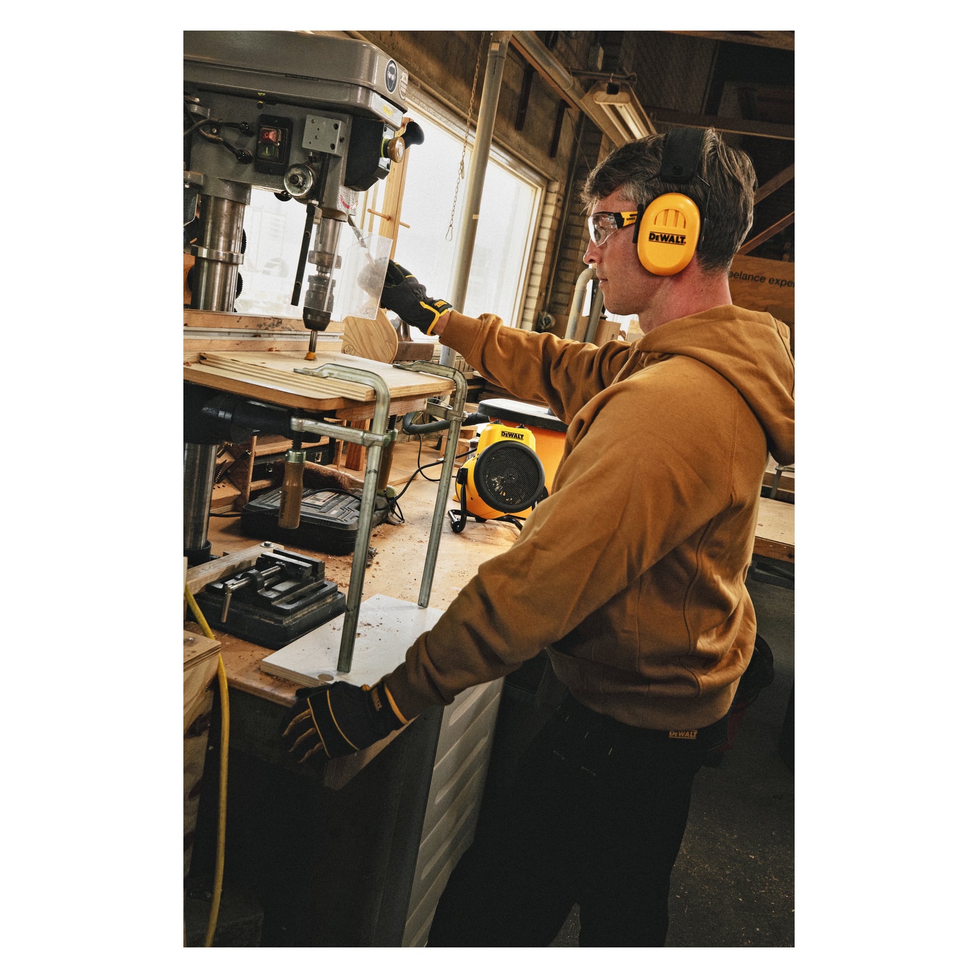 En person med skyddsglasögon, öronskydd, handskar och en brun hoodie använder en pelarborrmaskin i en verkstad. Arbetsbänken rymmer träbitar och verktyg, med en DeWalt Heater-fläkt DXJHXA0002E synlig i bakgrunden.