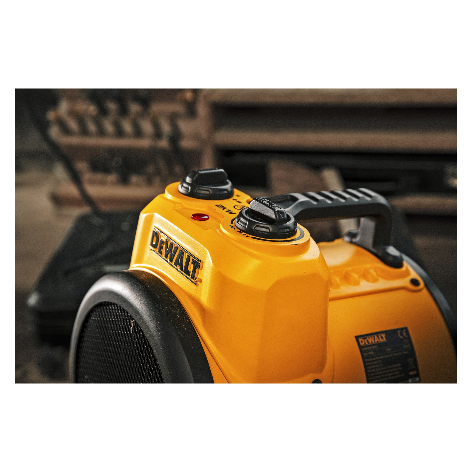 Närbild på en gul DeWalt Heater Fan DXJHXA0002E med två svarta styrknoppar och ett handtag ovanpå, placerad inomhus mot en suddig bakgrund av verktyg och utrustning.