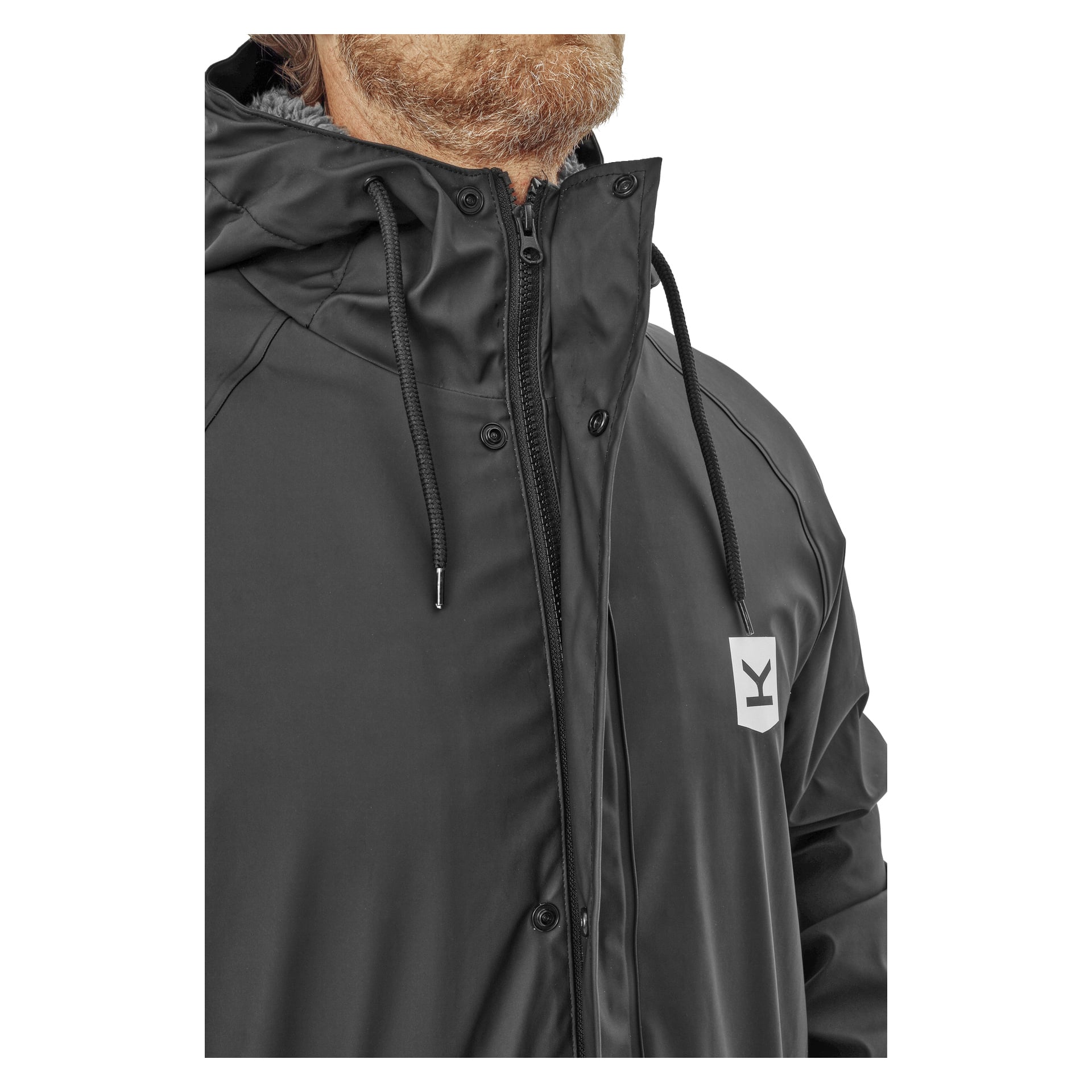 Eine Nahaufnahme zeigt eine Person in der schwarzen Kayoba Outdoor Men's Visby Regenjacke, die eine Kapuze mit Kordeln, einen Reißverschluss vorne und ein kleines weißes quadratisches Logo mit einem stilisierten K auf der Brust hat. Die Jacke ist teilweise geöffnet, um ihr graues Fleecefutter zu zeigen.