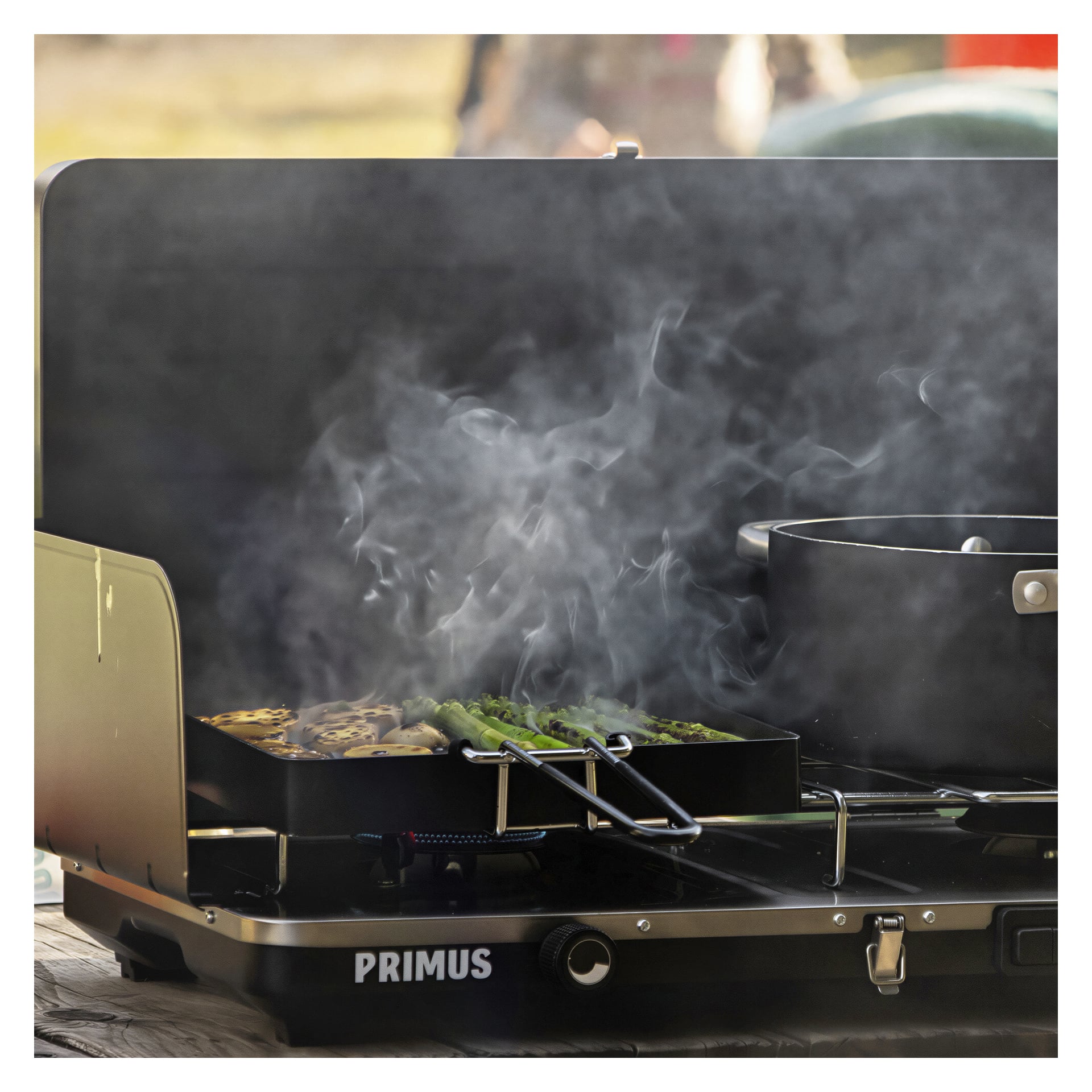 Primus Wilderness Grill Moto, med gryte og grillbrett, brukes utendørs til å tilberede grønnsaker som asparges og squash, med synlig damp eller røyk som stiger opp fra maten.
