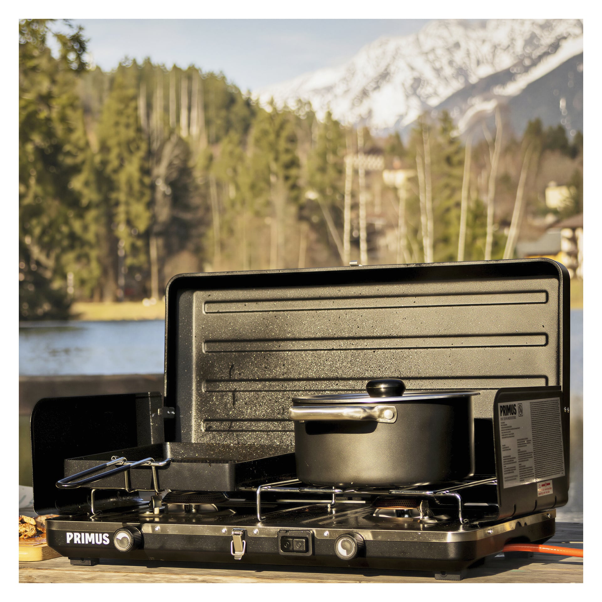 En Primus Wilderness-grill Moto med doble plater står på et trebord utenfor og lager mat i en svart gryte og panne. Bak den ligger snødekte fjell, trær og en innsjø.