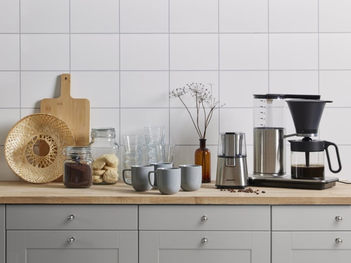 Eine Küchentheke zeigt eine Kaffeemaschine, eine Mühle, Tassen, Glasgefäße mit Keksen und Kaffee, klare Gläser, einen geflochtenen Korb, ein Holzschneidebrett und eine braune Vase mit getrockneten Blumen vor einer gefliesten Wand.