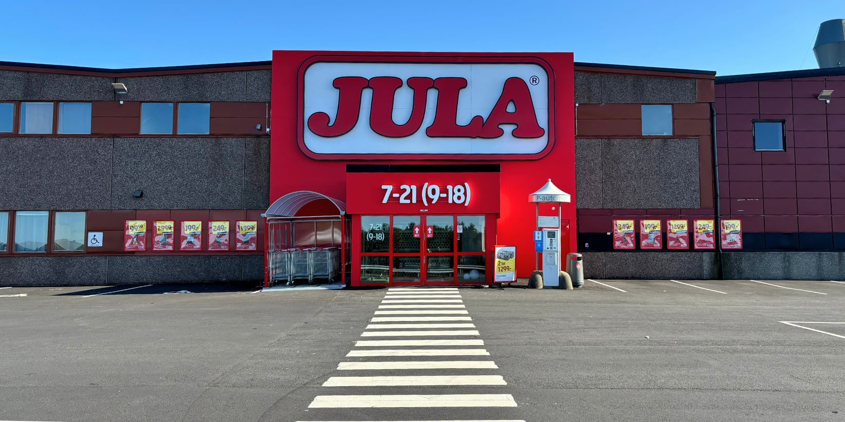 En romslig Jula-butikk med en rød inngang, glassdører og rekker av handlekurver utenfor. Et fotgjengerfelt leder besøkende til inngangen, og åpningstidene er oppslått over dørene. Parkeringsplassen er tom.