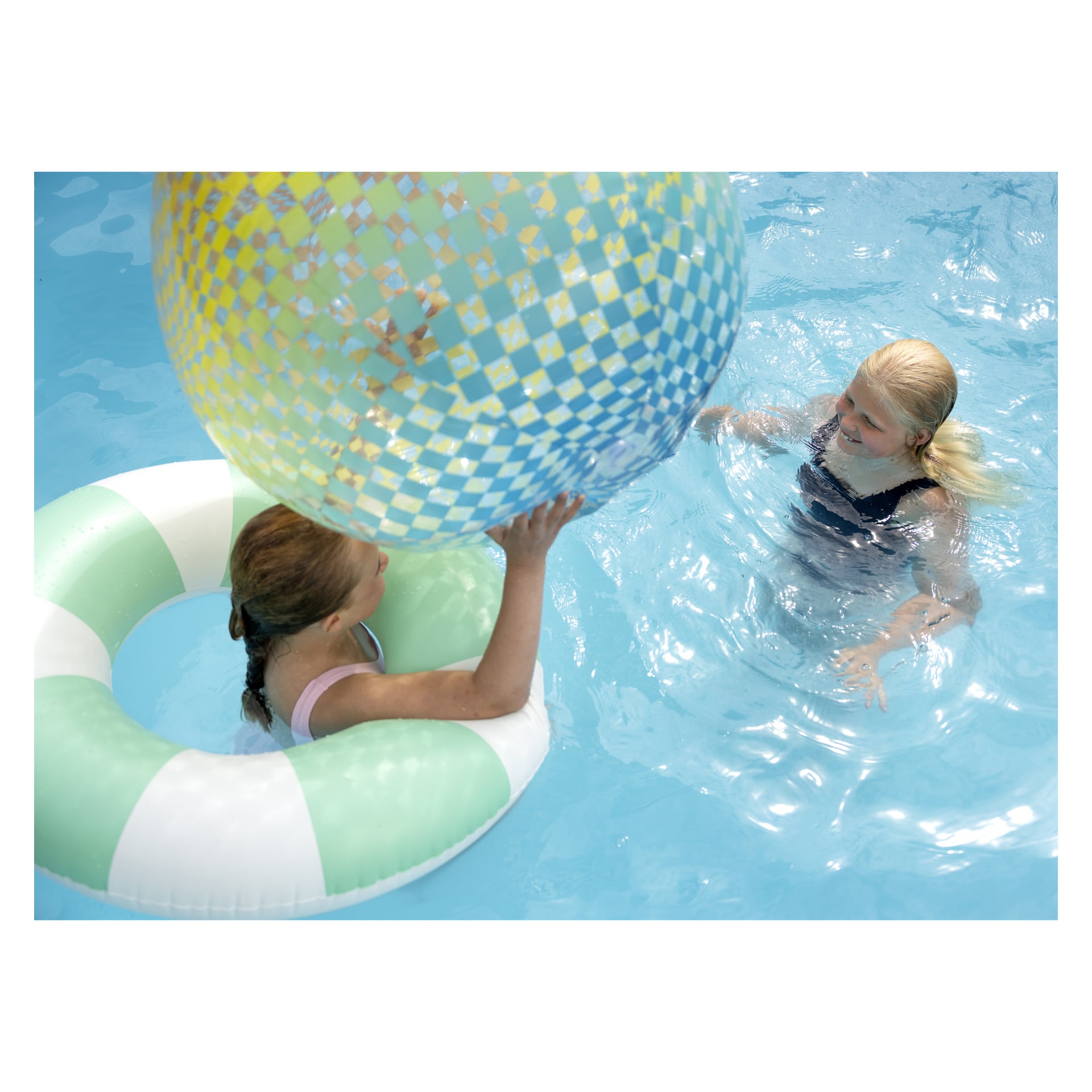 Kaksi lasta leikkii uima-altaassa, jossa on suuri ruudullinen Colosphere Beach Ball Bestwaylta ja vihreä-valkoraidallinen ilmatäytteinen uimarengas.