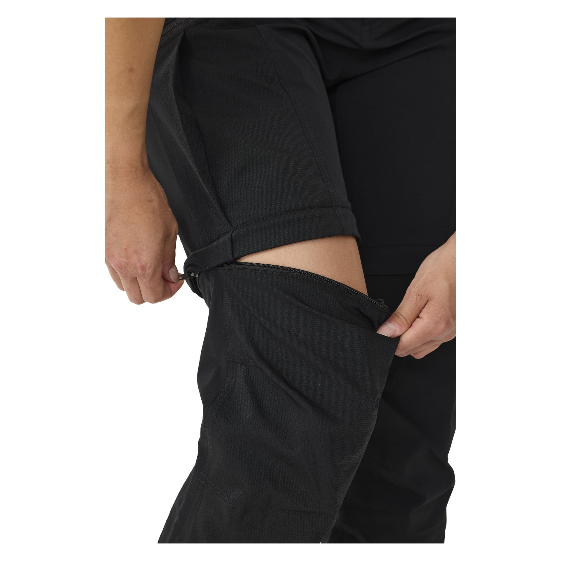 En hånd glir ned den nedre delen av Kayoba Outdoors Women's Abisko utendørsbukser med glidelåsbena, og gjør dem om til shorts. Buksene er delvis åpne over kneet, noe som eksponerer noe hud.