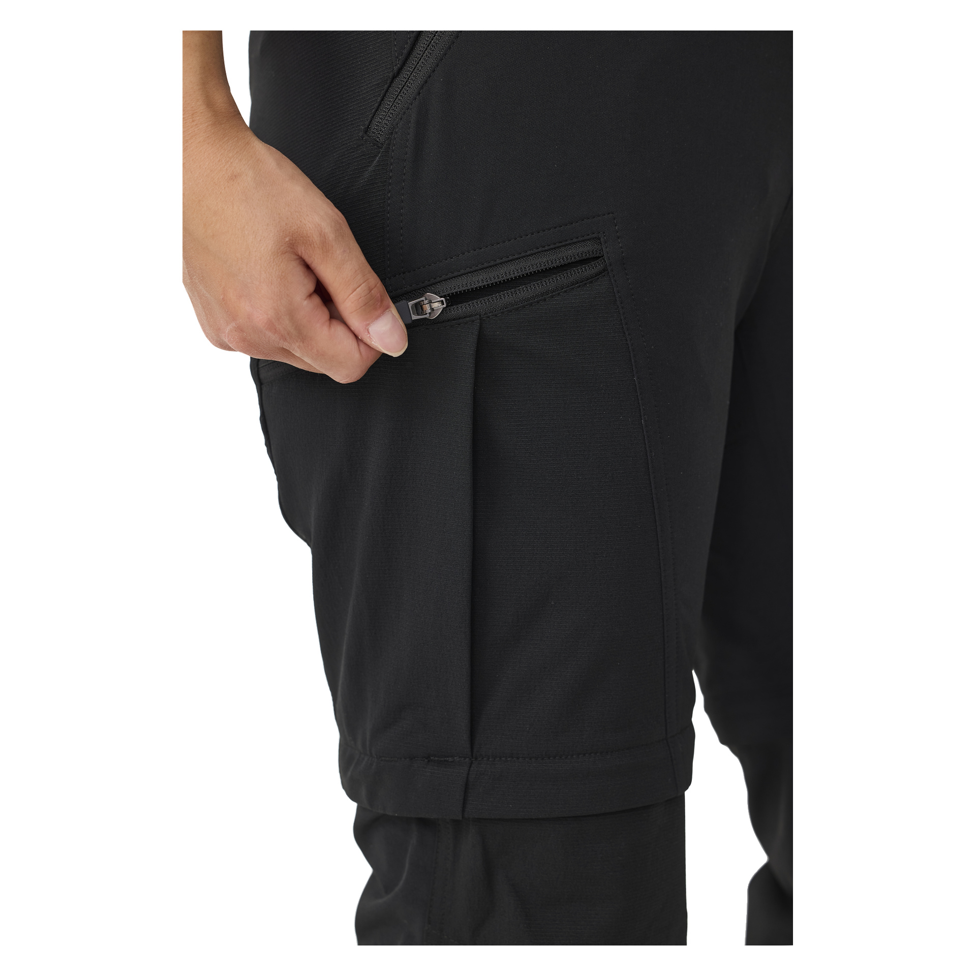 En hand greppar dragkedjan på en svart ficka på Kayoba Outdoor Women's utomhusbyxor med dragkedje-ben, som visar delar av bärarens ben och hand. Byxorna är gjorda av slät tyg och har en framträdande ficka med dragkedja.