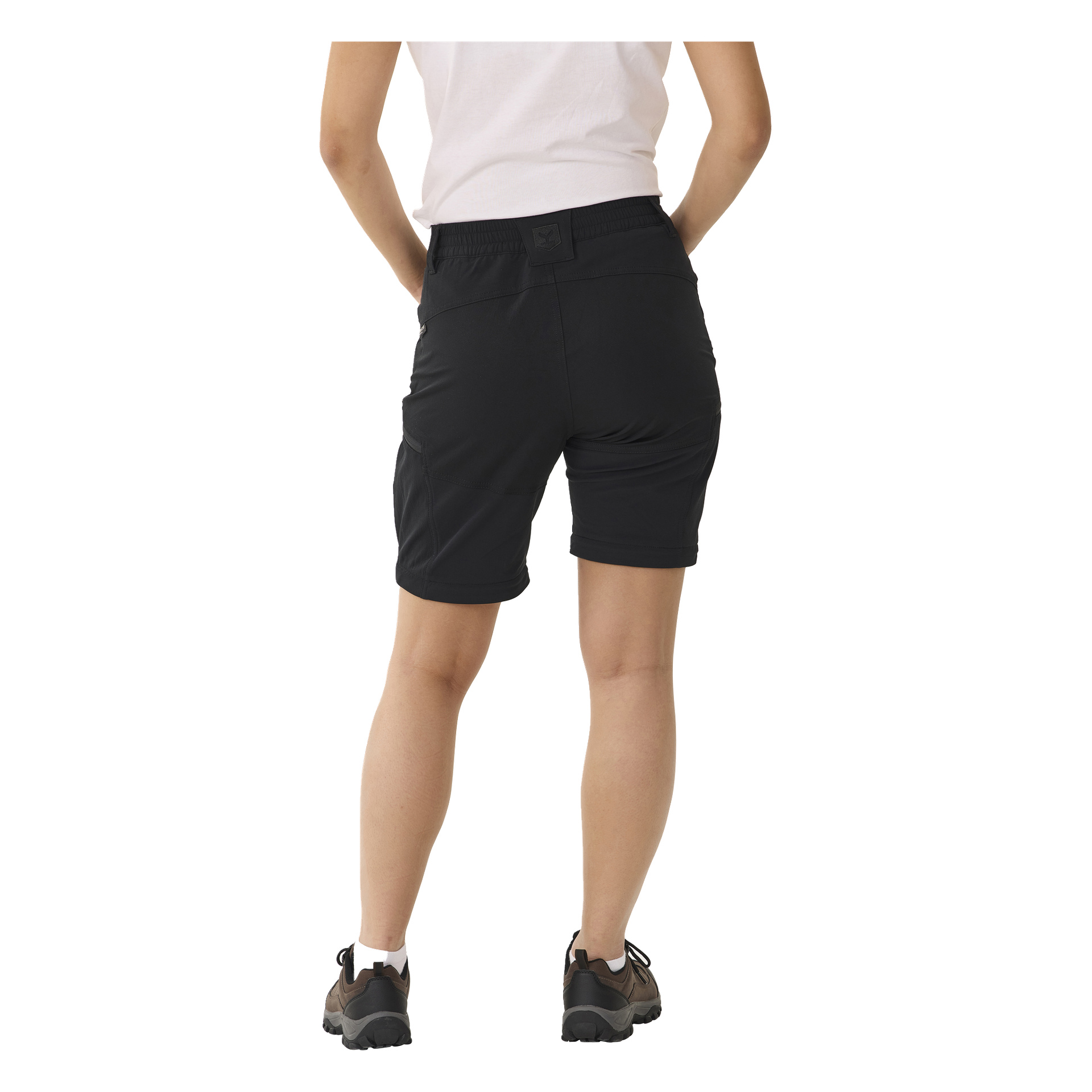 En person står med ryggen mot kameran, klädd i en vit topp, svarta Kayoba Outdoor Women's byxor med dragkedje-ben Abisko och svarta vandringsskor. Deras händer är placerade bakom ryggen.