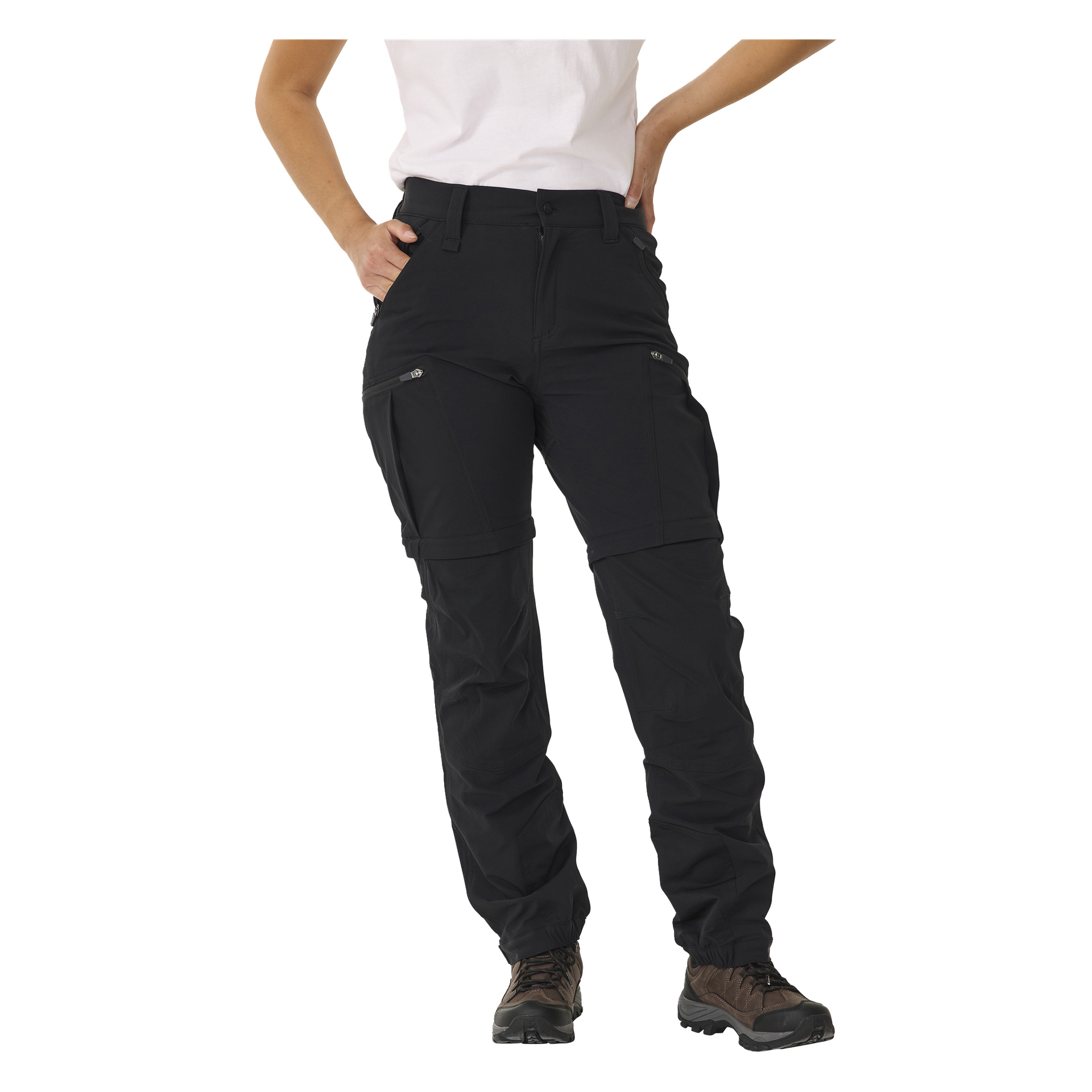 Någon står med ena handen vilande på höften, klädd i svarta Kayoba Outdoor Women's zip-off-byxor med dragkedjefickor framtill och på sidorna. Outfiten kompletteras med en vit kortärmad skjorta och bruna vandringsskor.