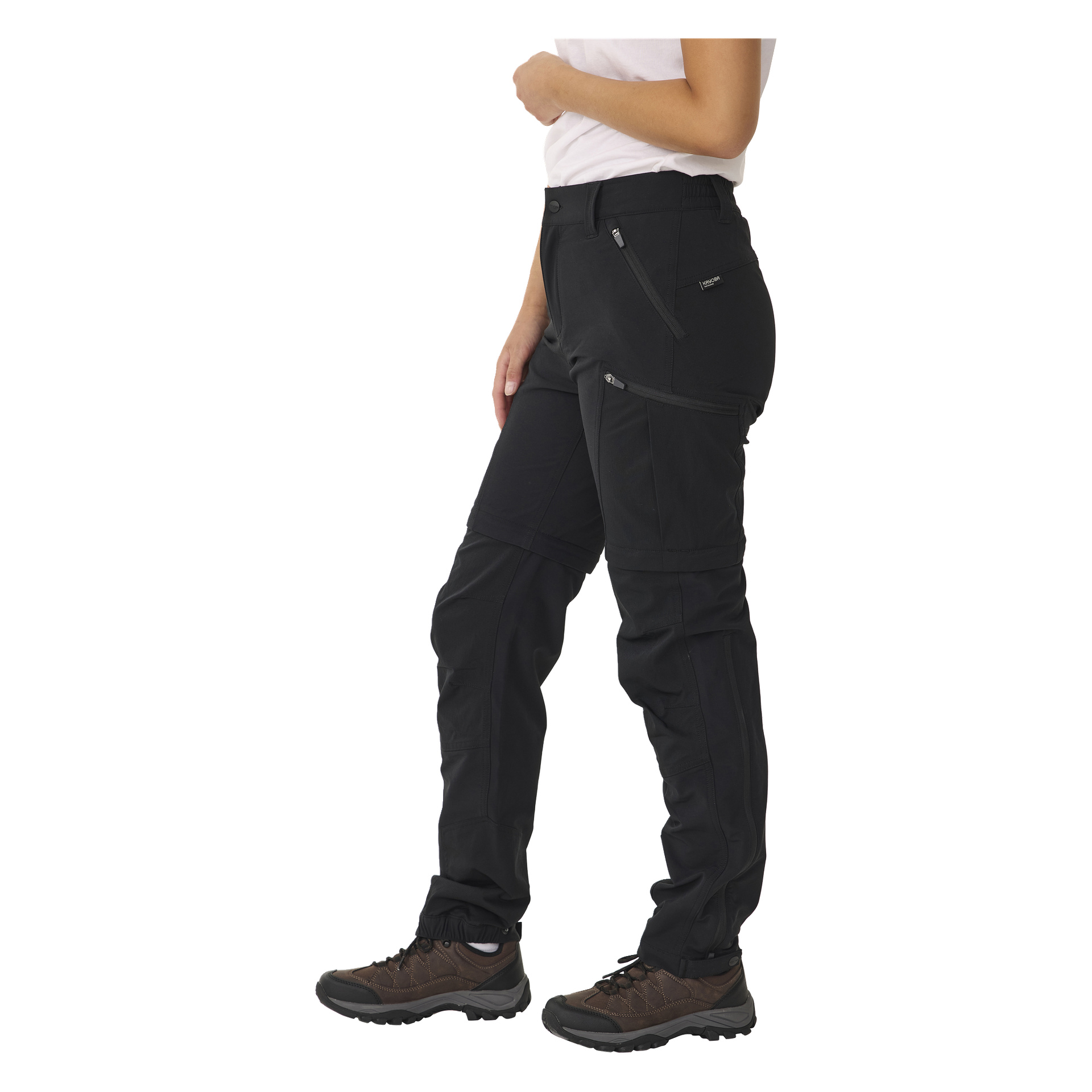 En person är klädd i en vit kortärmad skjorta, bruna vandringsskor och svarta Kayoba Outdoor Women's utomhusbyxor med dragkedjeben. Byxorna har flera fickor och dragkedjor.