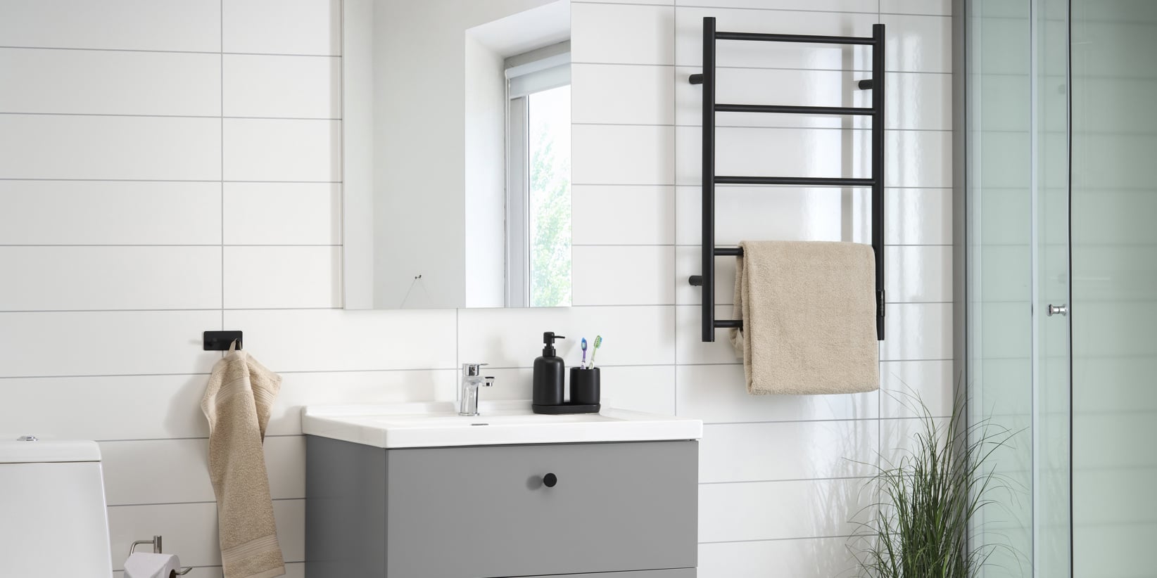 Modernes Badezimmer mit weißen Fliesenwänden, einem grauen Waschbecken mit weißem Waschbecken, einem schwarzen Handtuchhalter mit beigem Handtuch, einem Spiegel über dem Waschbecken, Seifenspendern, einem Fenster, das das Licht hereinlässt, einer Glasduschtür und einer Topfpflanze, die Grün spendet.